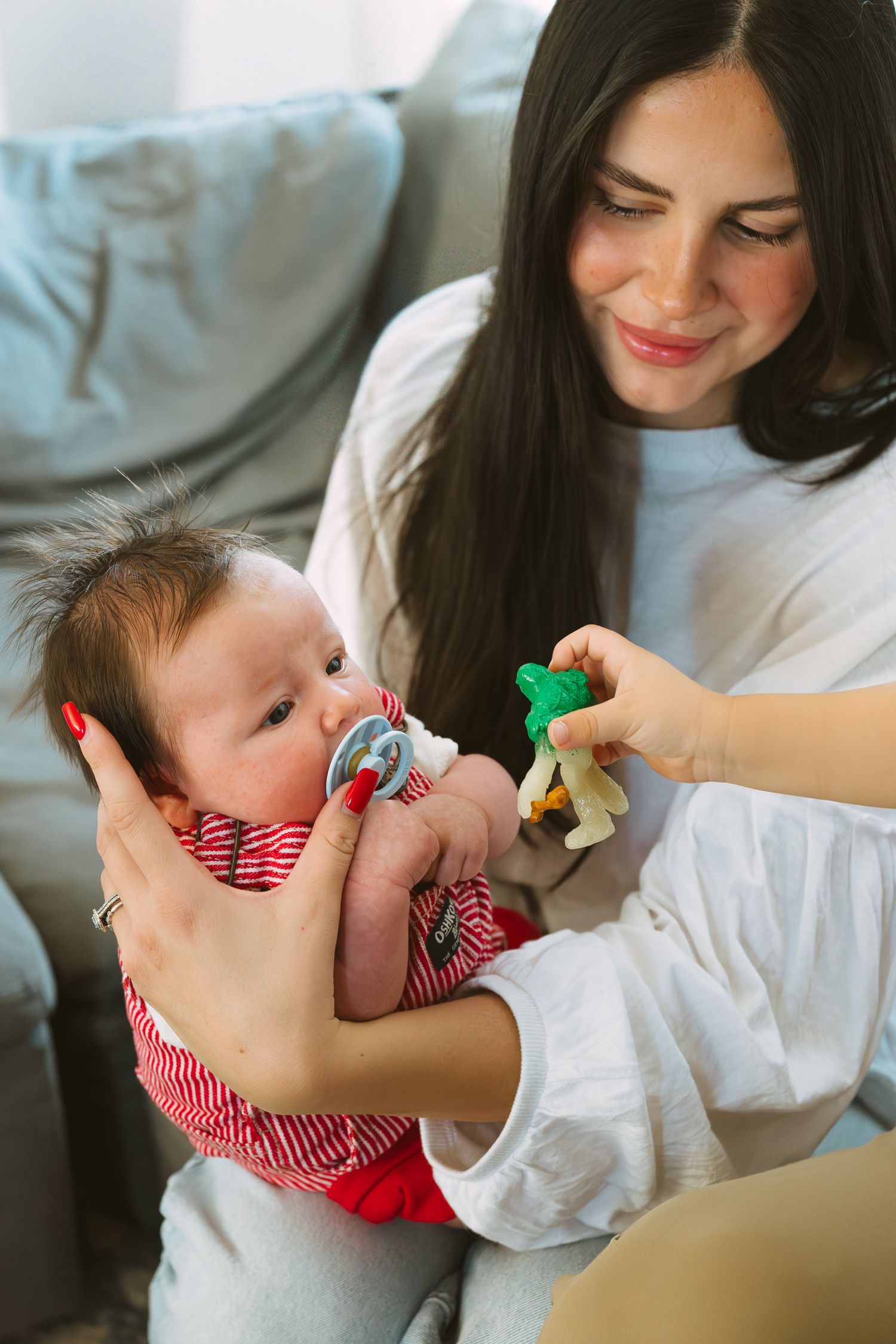A tender moment on a couch as a baby in red stripes plays with a green toy while being cradled.