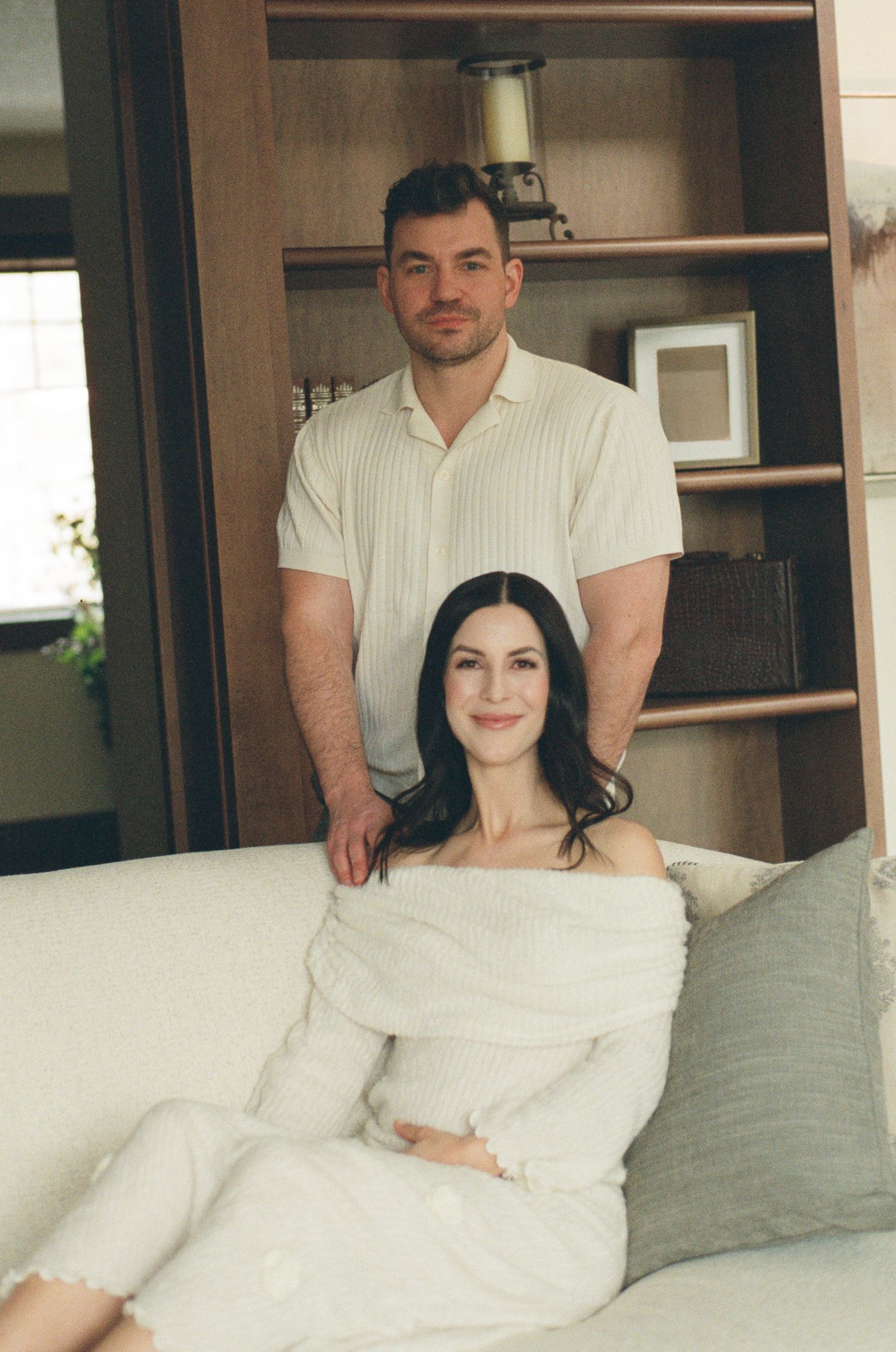 Couple poses indoors, man standing behind pregnant woman in white off-shoulder dress on couch.