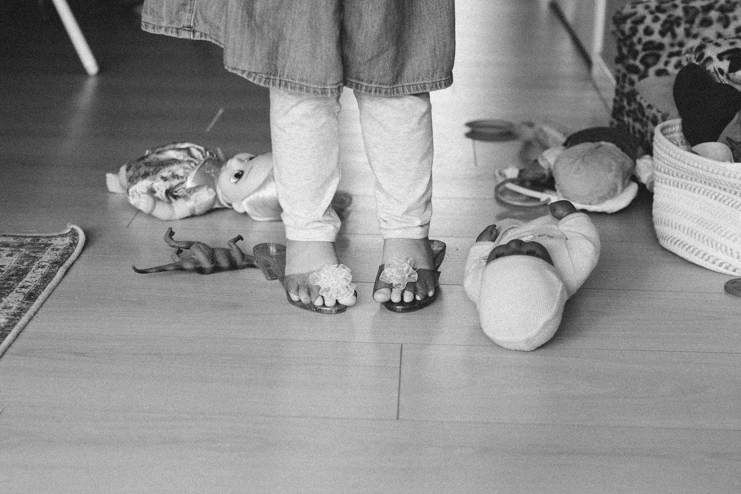 Black and white photo of a child standing on hardwood floor surrounded by stuffed animal toys wearing sandals.