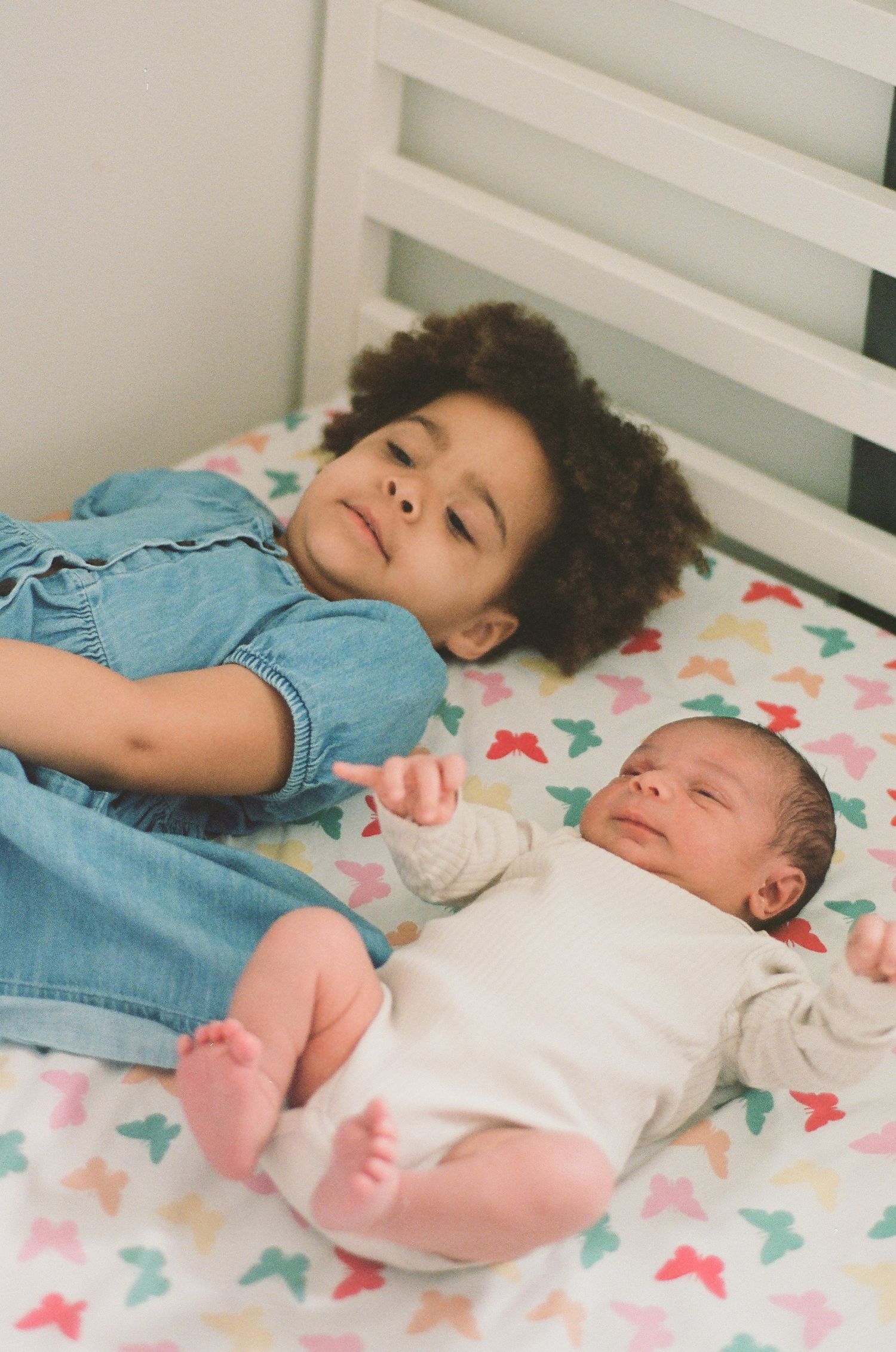 Young girl with curly hair lies on patterned bed beside newborn baby, gently pointing at the infant.
