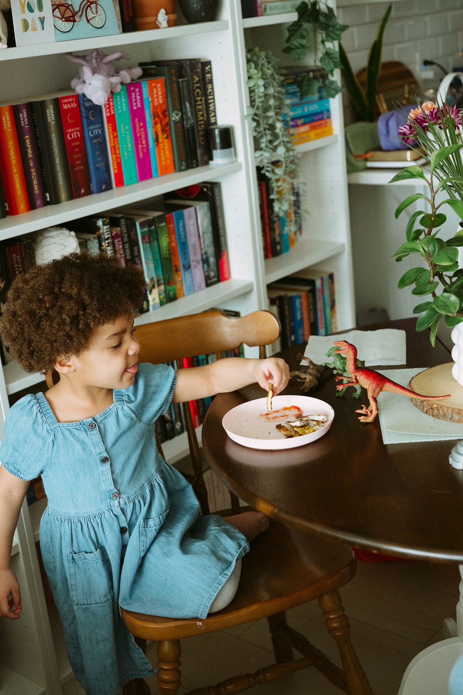 Young girl in denim dress eating at wooden table with dinosaur toys, bookshelf in background.