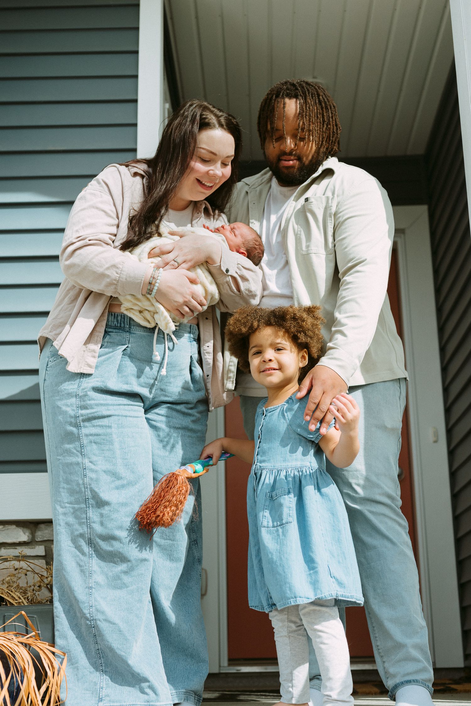 Happy interracial family of four posing on home steps, mother holding newborn baby, father beside young curly-haired daughter.