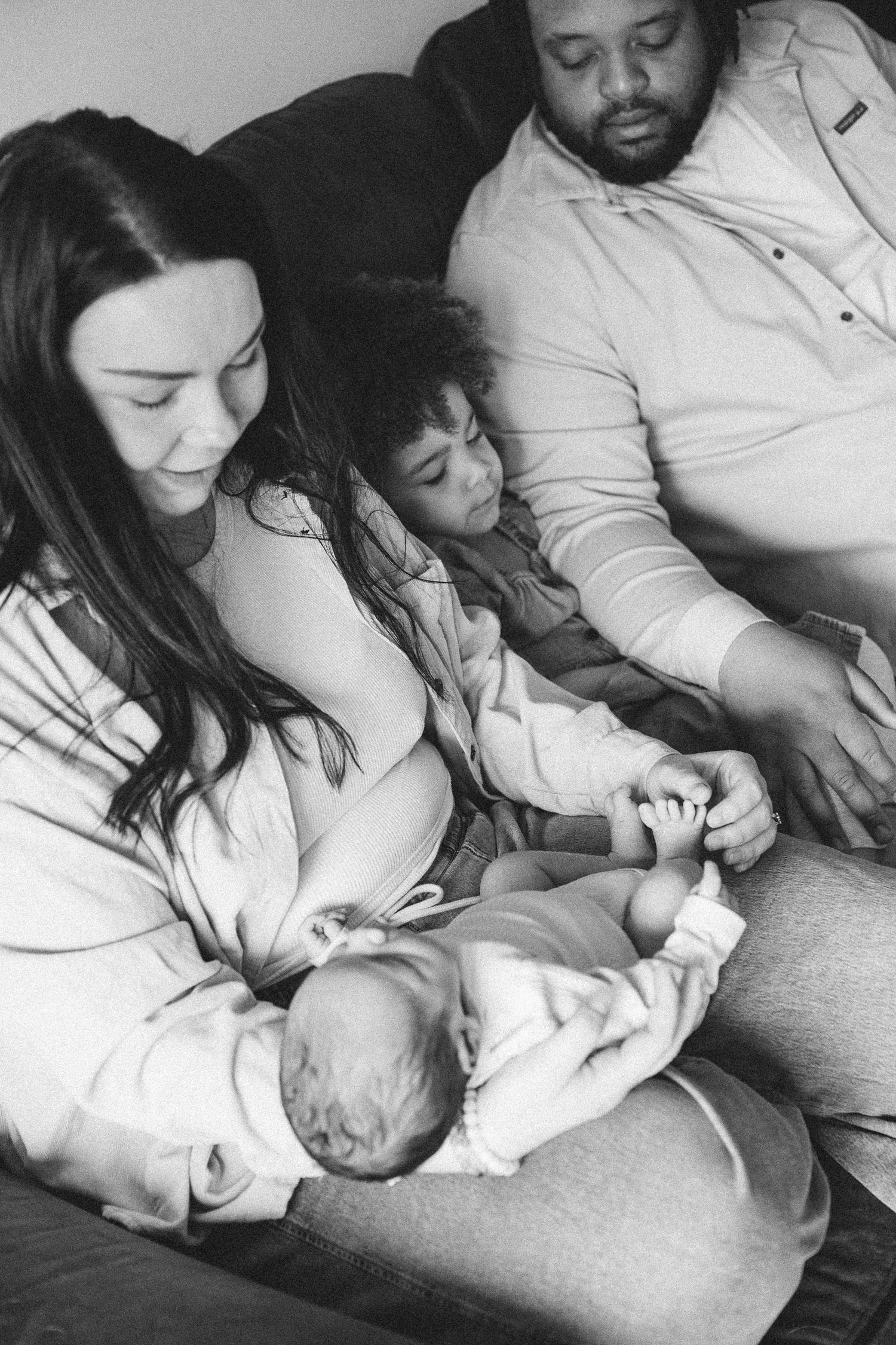 Black and white family photo of parents and toddler admiring their newborn baby on a couch.