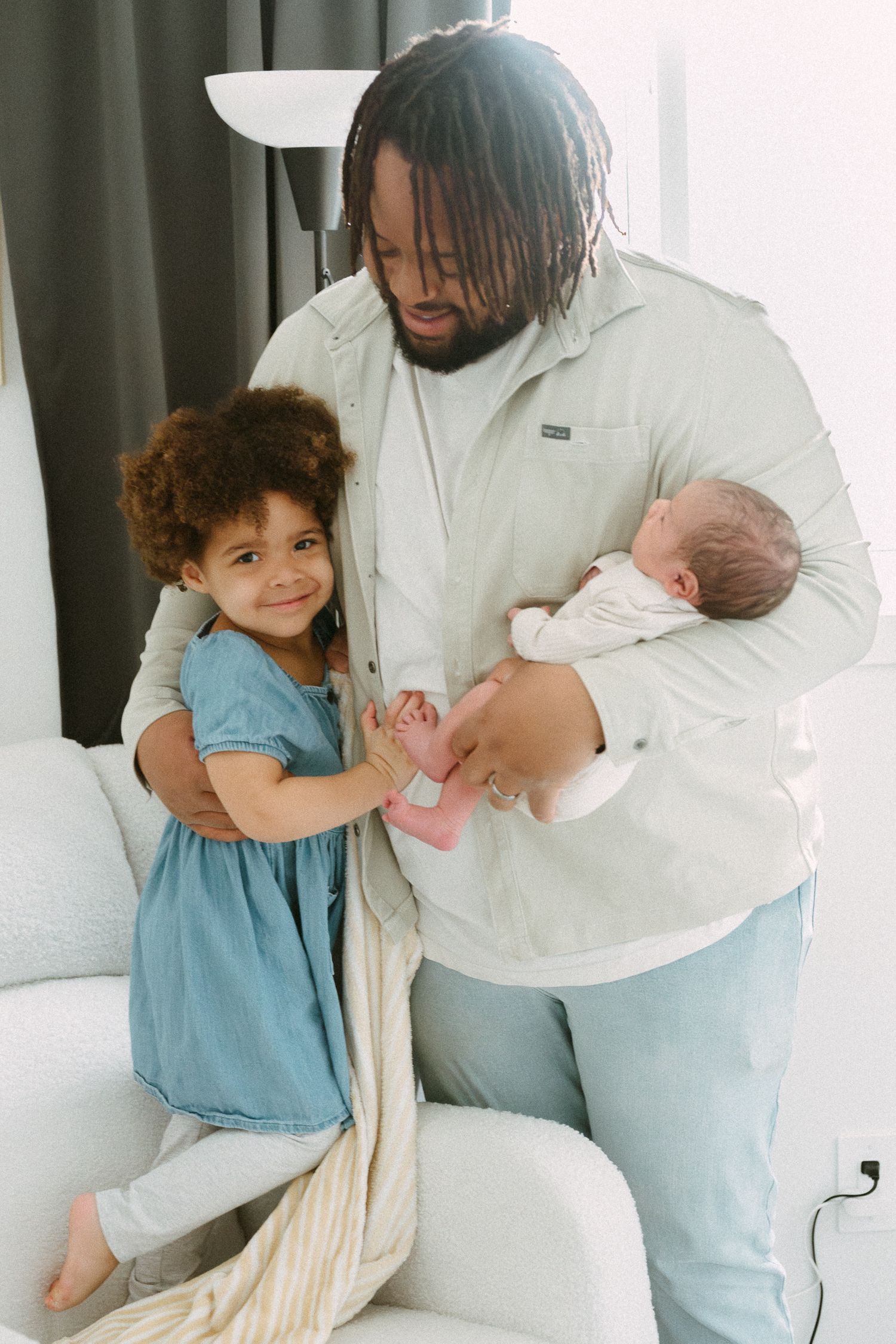 Father in sage jacket holds newborn as curly-haired toddler girl smiles and touches baby's hand indoors.