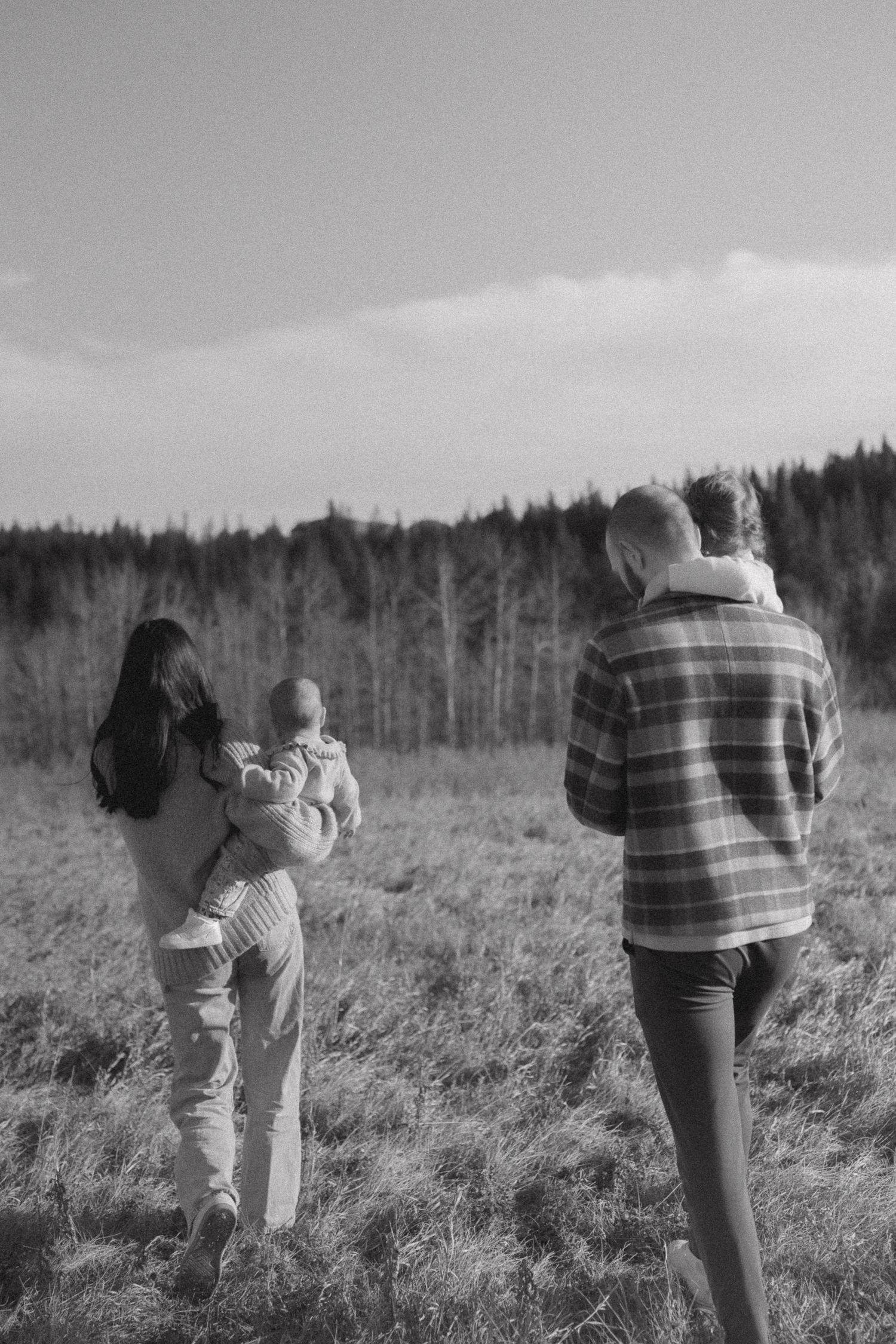 Family walking together through a grassy field with evergreen trees in the background on a cloudy day.