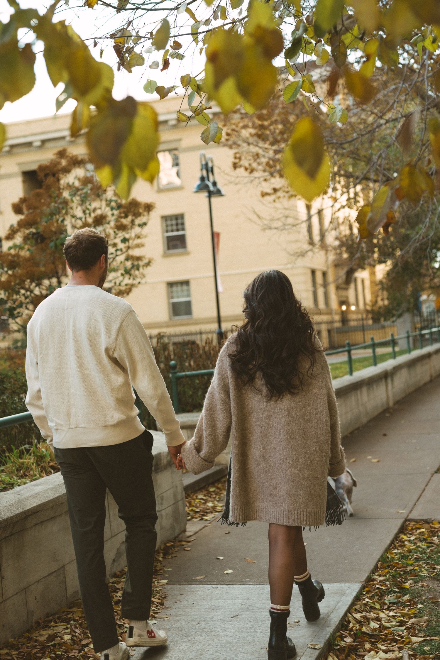 Two people walk together on a path lined with autumn leaves and stone railings on a sunny day.