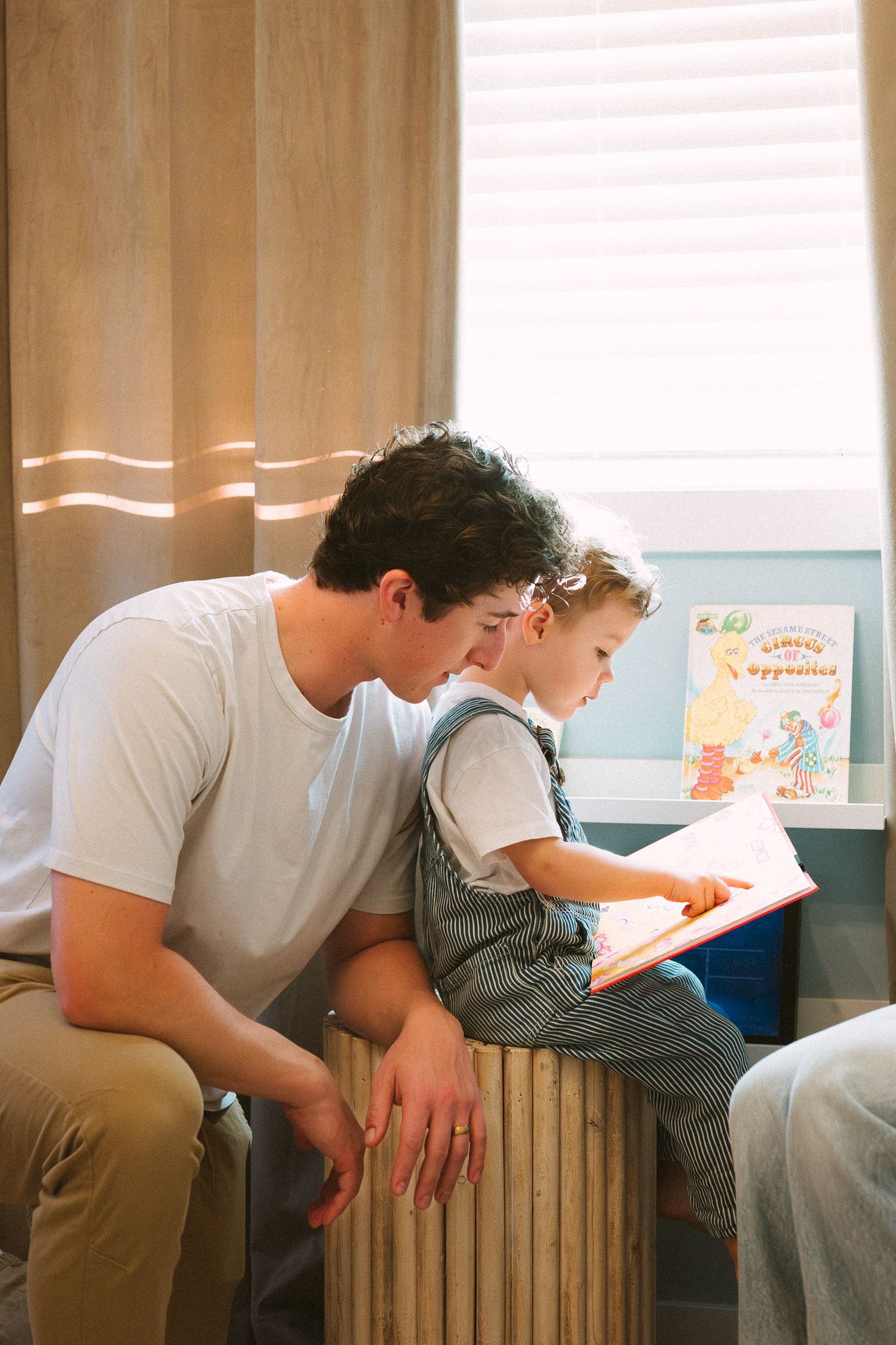 A parent and child sit together on a bench reading a book in a sunlit room.