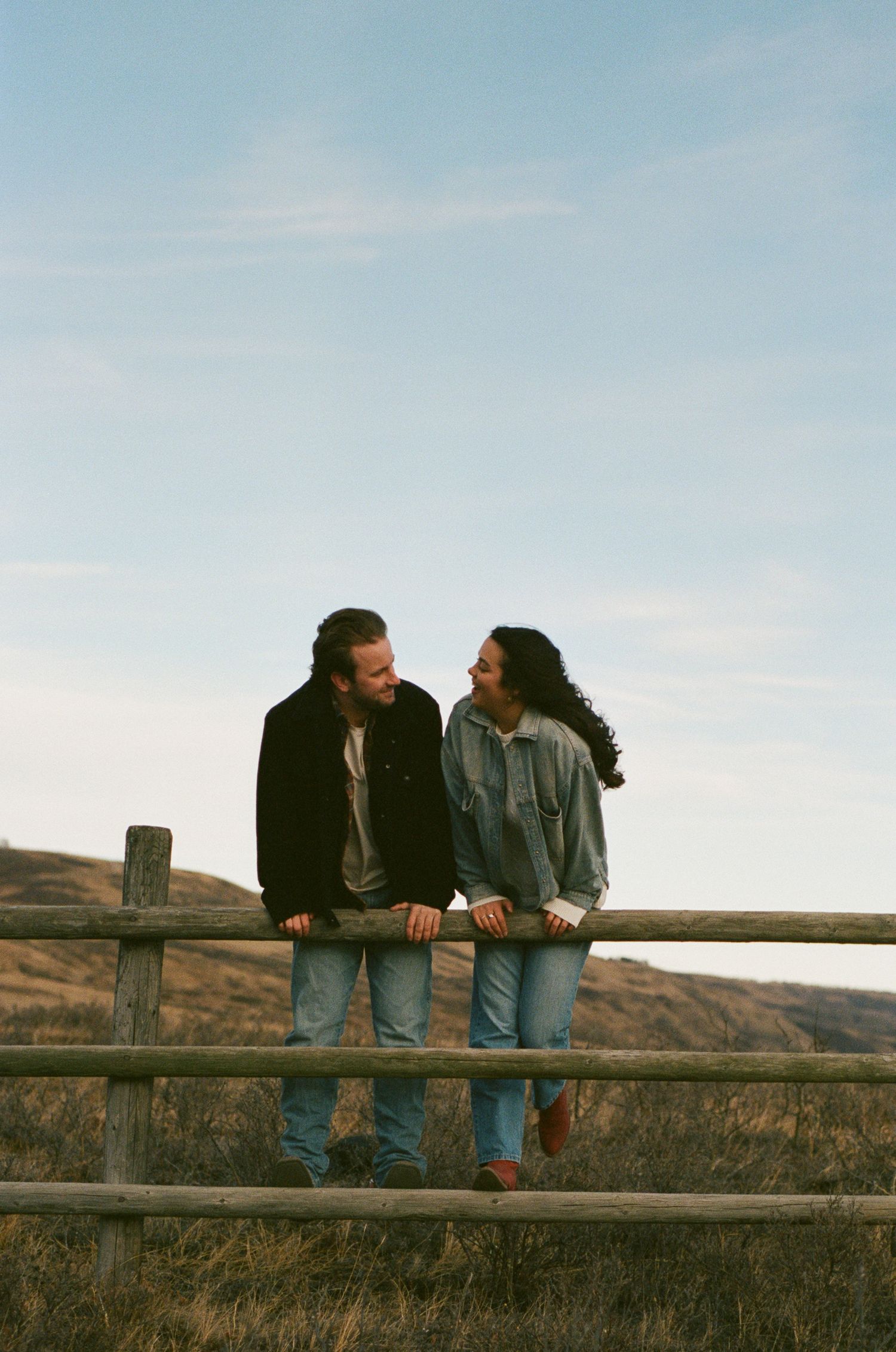 Two people lean against a rustic wooden fence with scenic hills and grassland in the background.