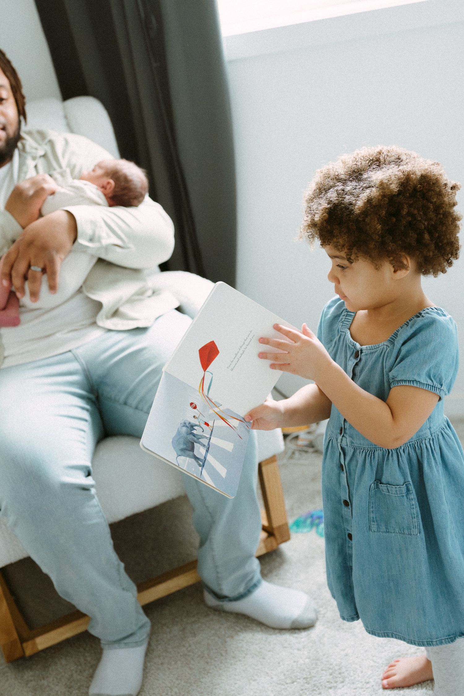 A father holds a newborn baby while his toddler daughter in a blue dress looks at a colorful picture book.