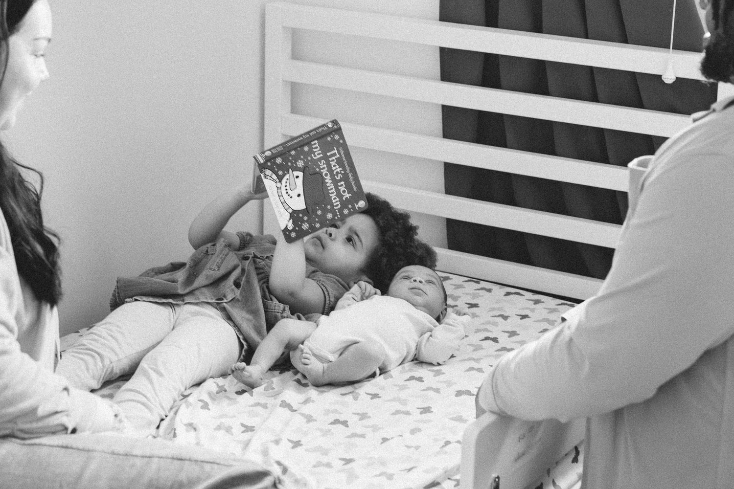 Black and white photo of a toddler lying on a bed reading a book next to a newborn baby with parents nearby.