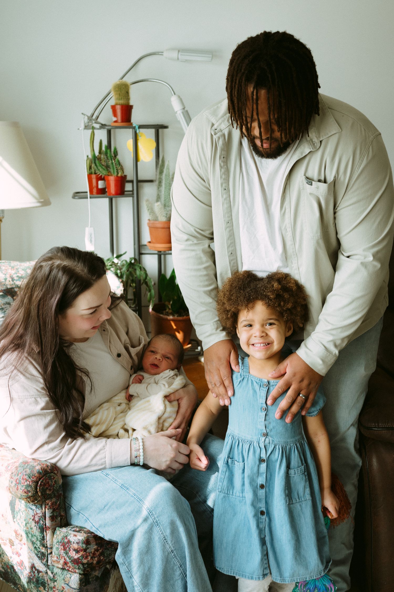 A happy interracial family of four poses together, with mother holding newborn as father stands beside smiling daughter.