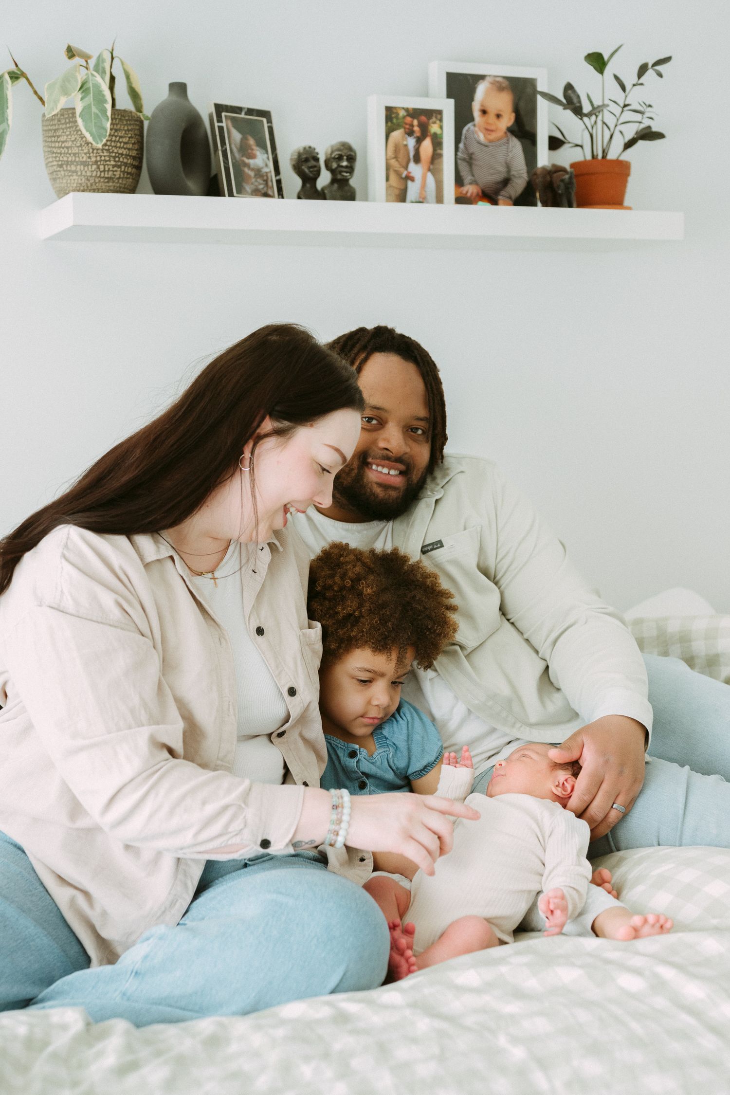 A happy interracial family sits on a bed, parents smiling at their newborn baby while their toddler looks on.