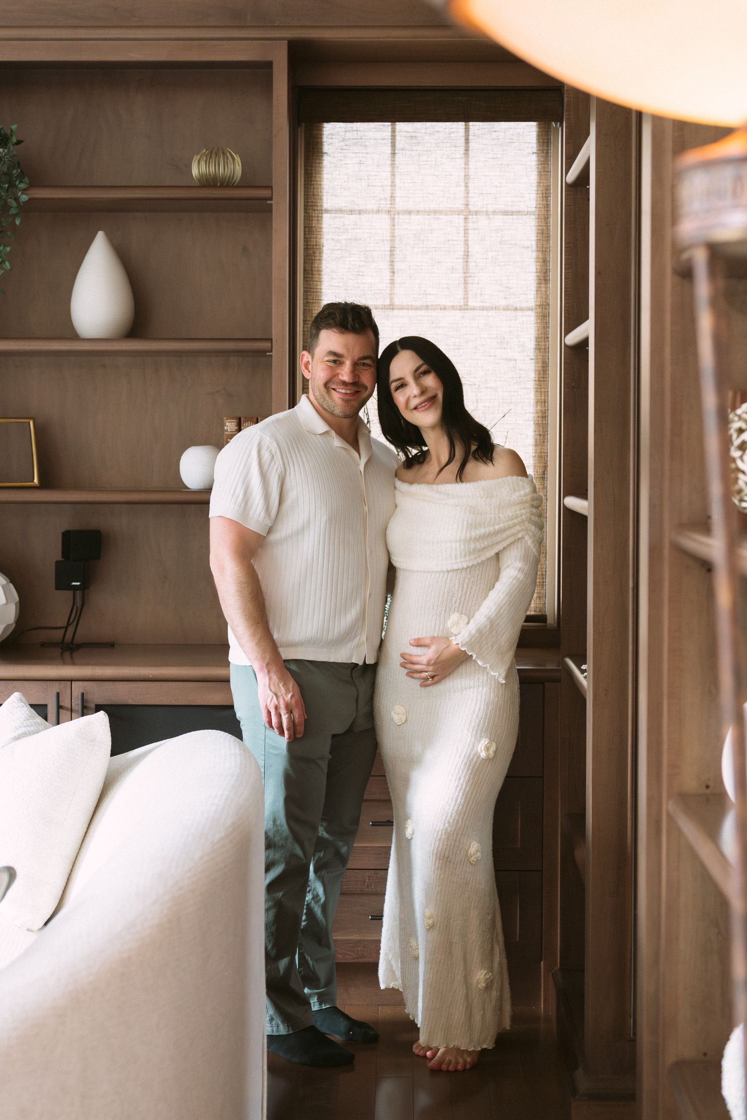 Smiling couple posing together, pregnant woman in white floral dress with partner in neutral outfit in cozy wood interior.