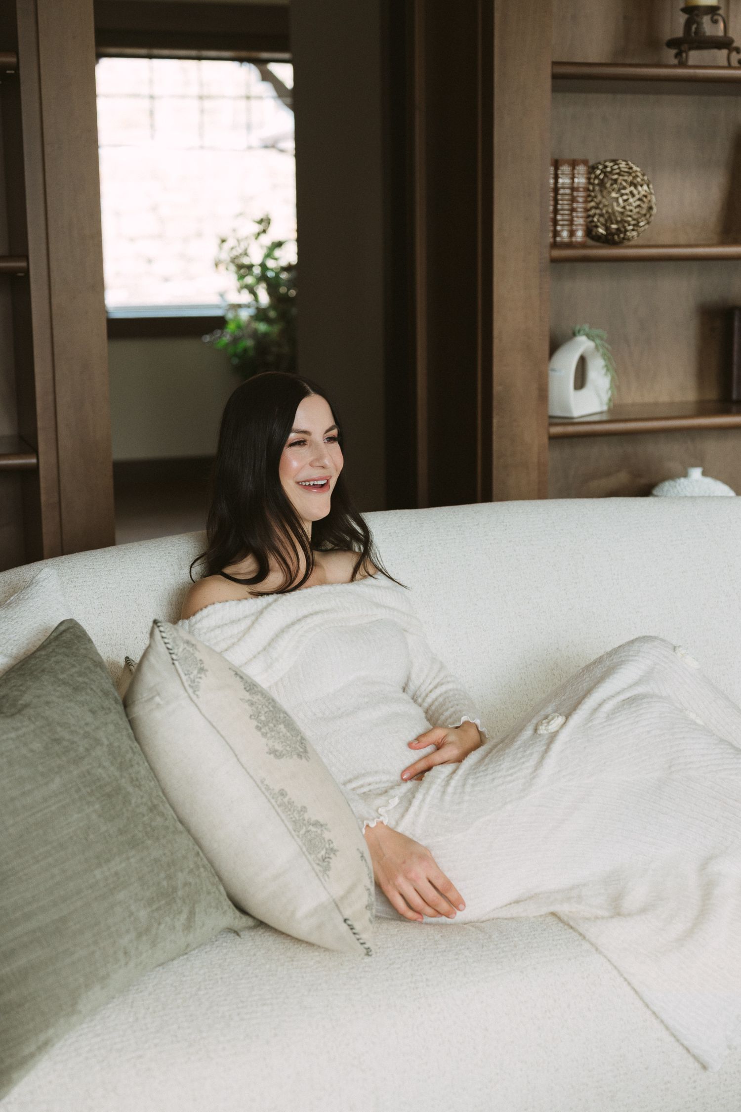 Laughing pregnant woman in white off-shoulder dress relaxing on cream sofa with pillows in cozy wood-paneled room.