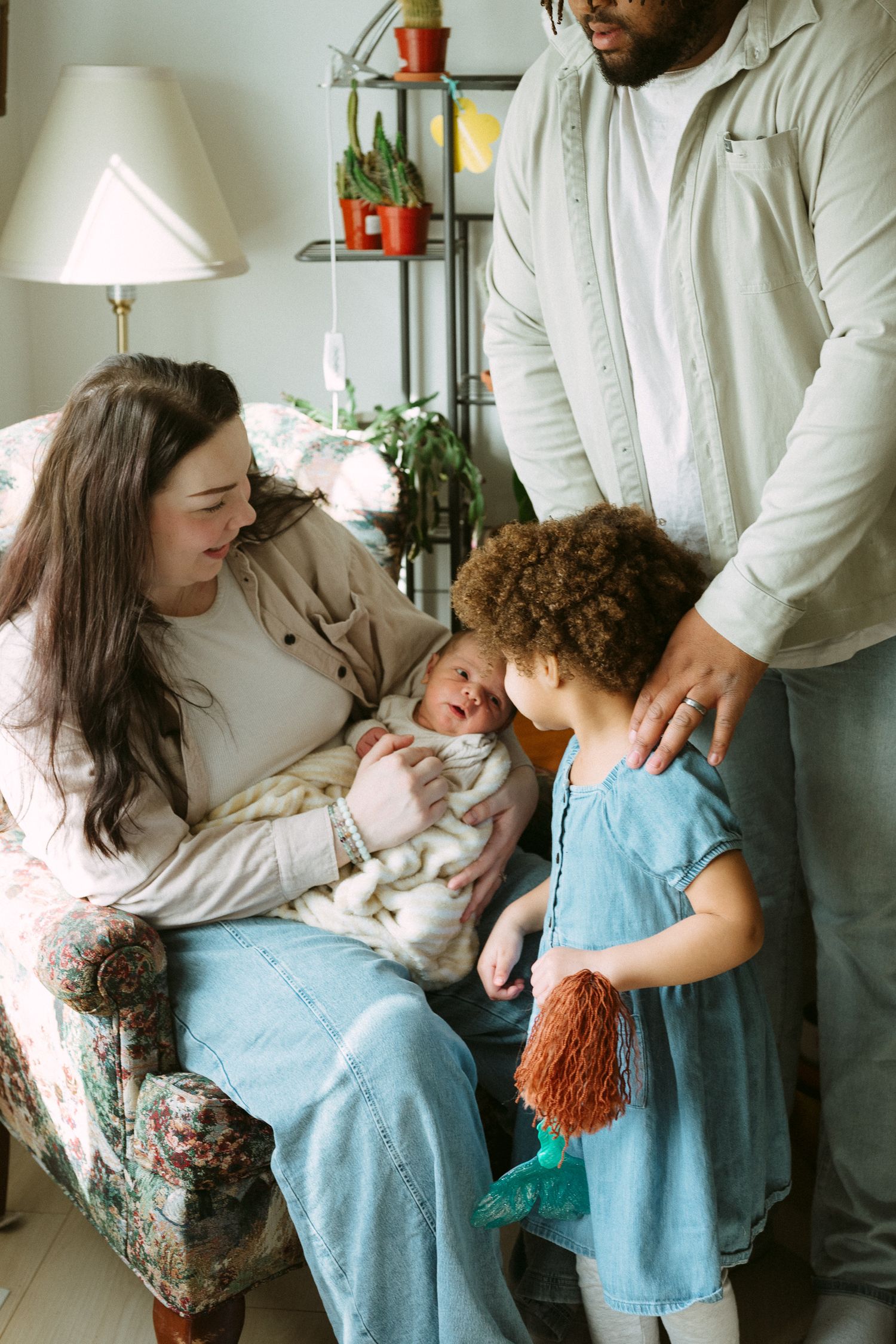 Family gathered around mother holding newborn baby, with young girl in blue dress and father standing nearby.