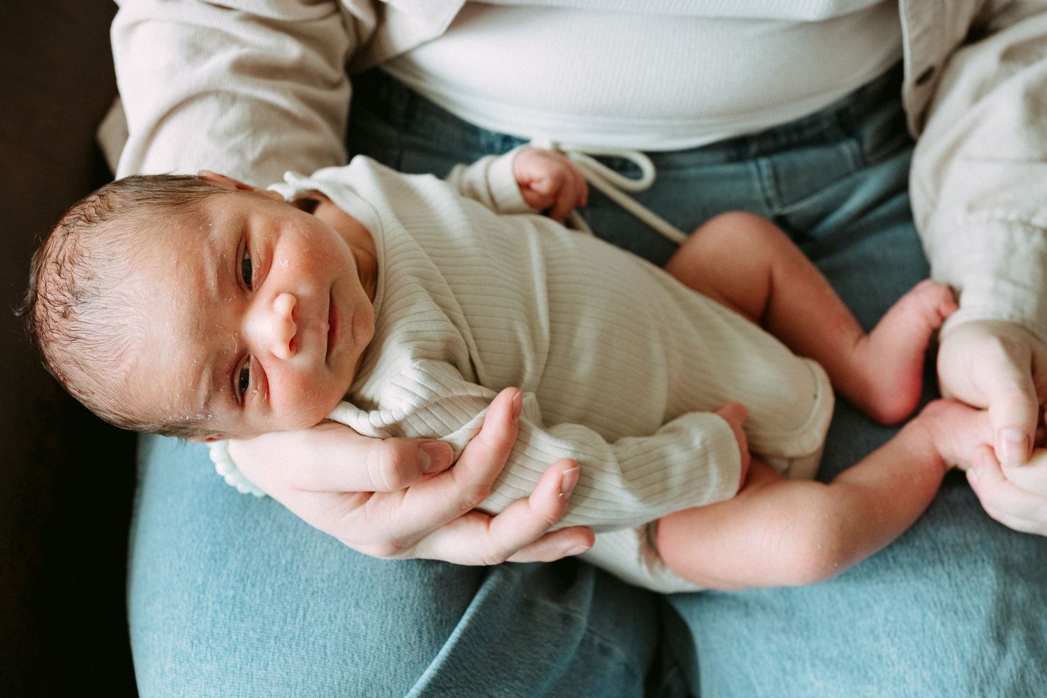 Newborn baby smiling while being cradled in adult hands, wearing a cream onesie, with tiny bare feet visible.