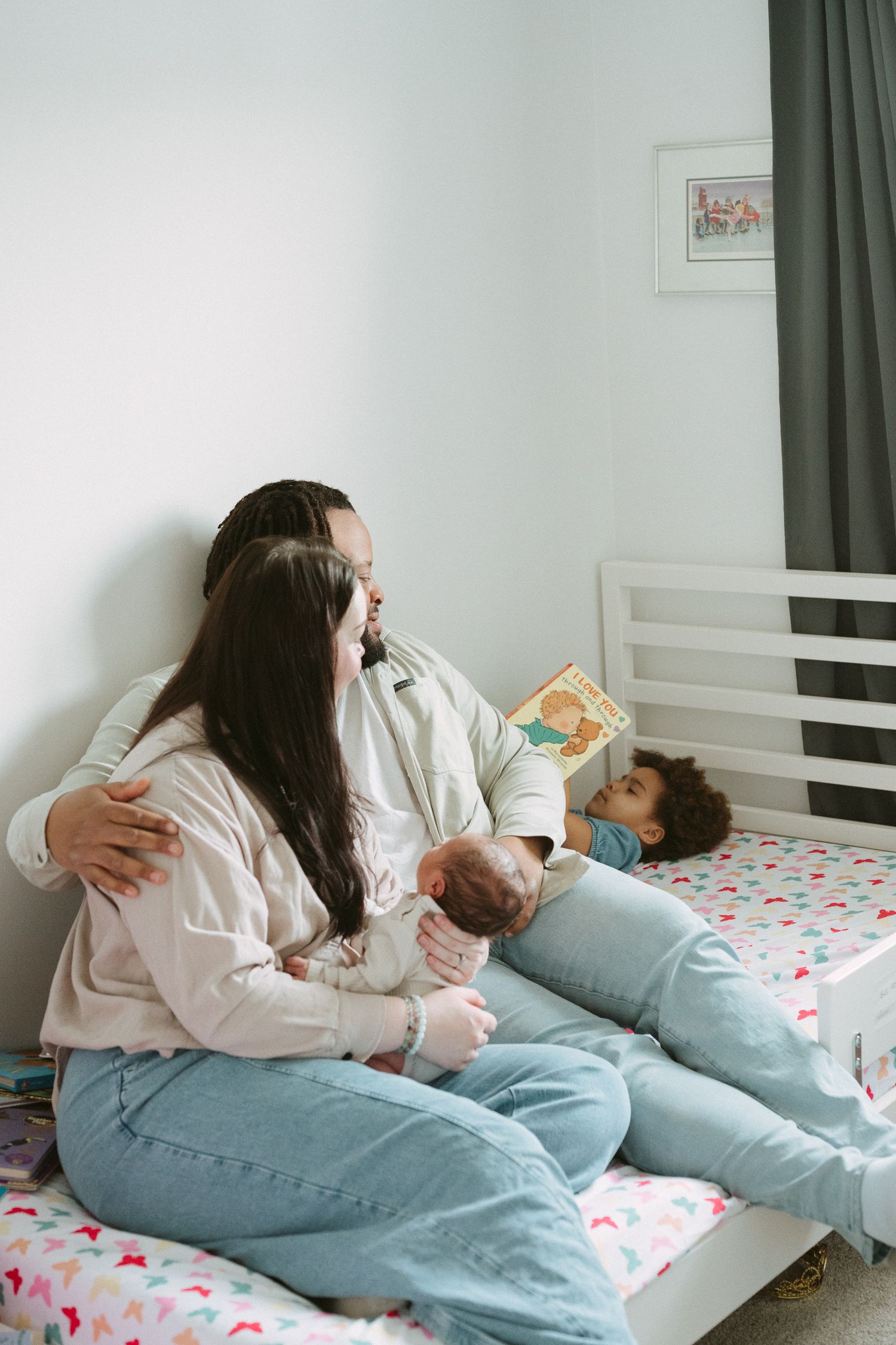 A mother sits on a child's bed holding a newborn while an older child reads a picture book beside her.