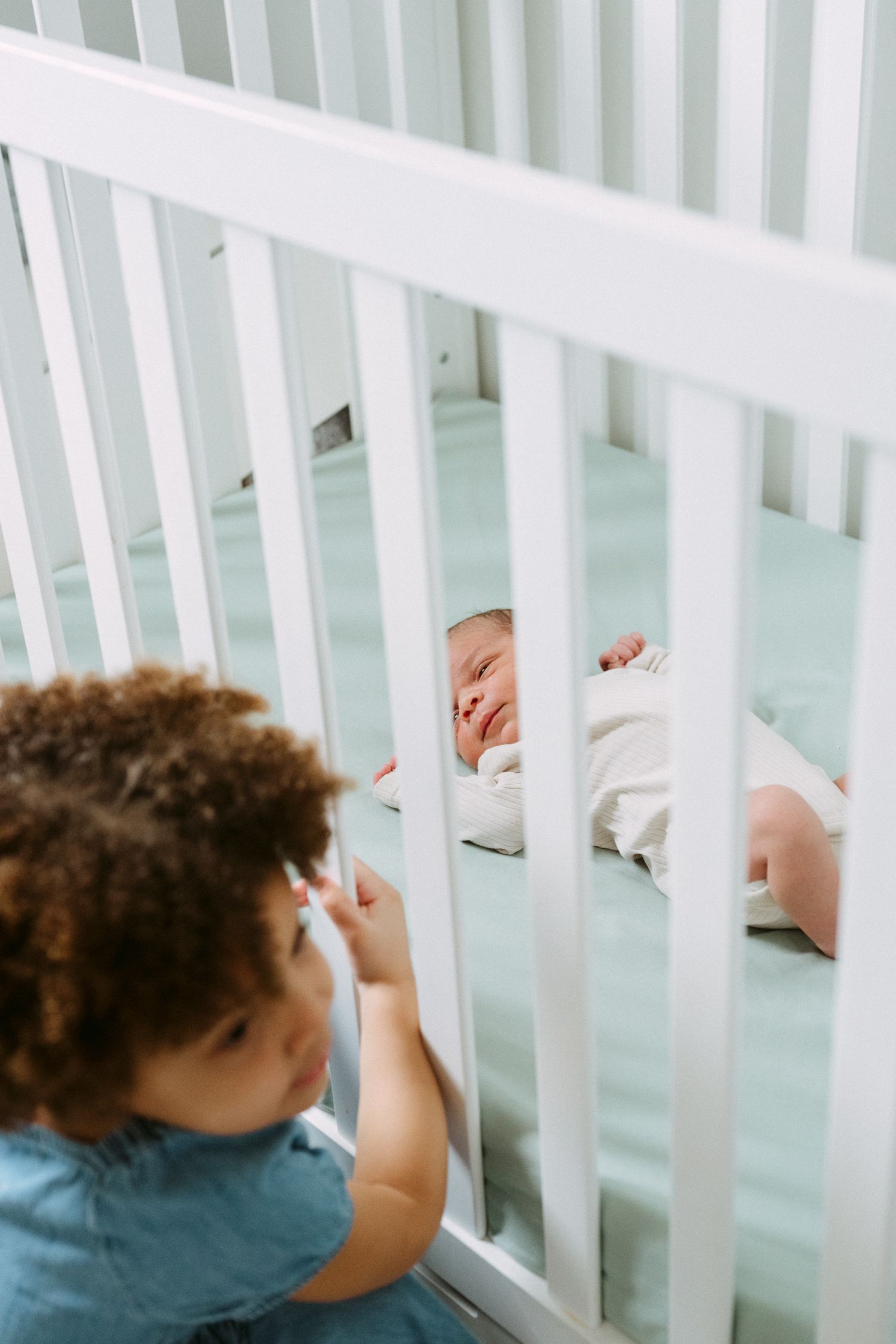Curly-haired toddler peering through white crib rails at sleeping newborn baby sibling.
