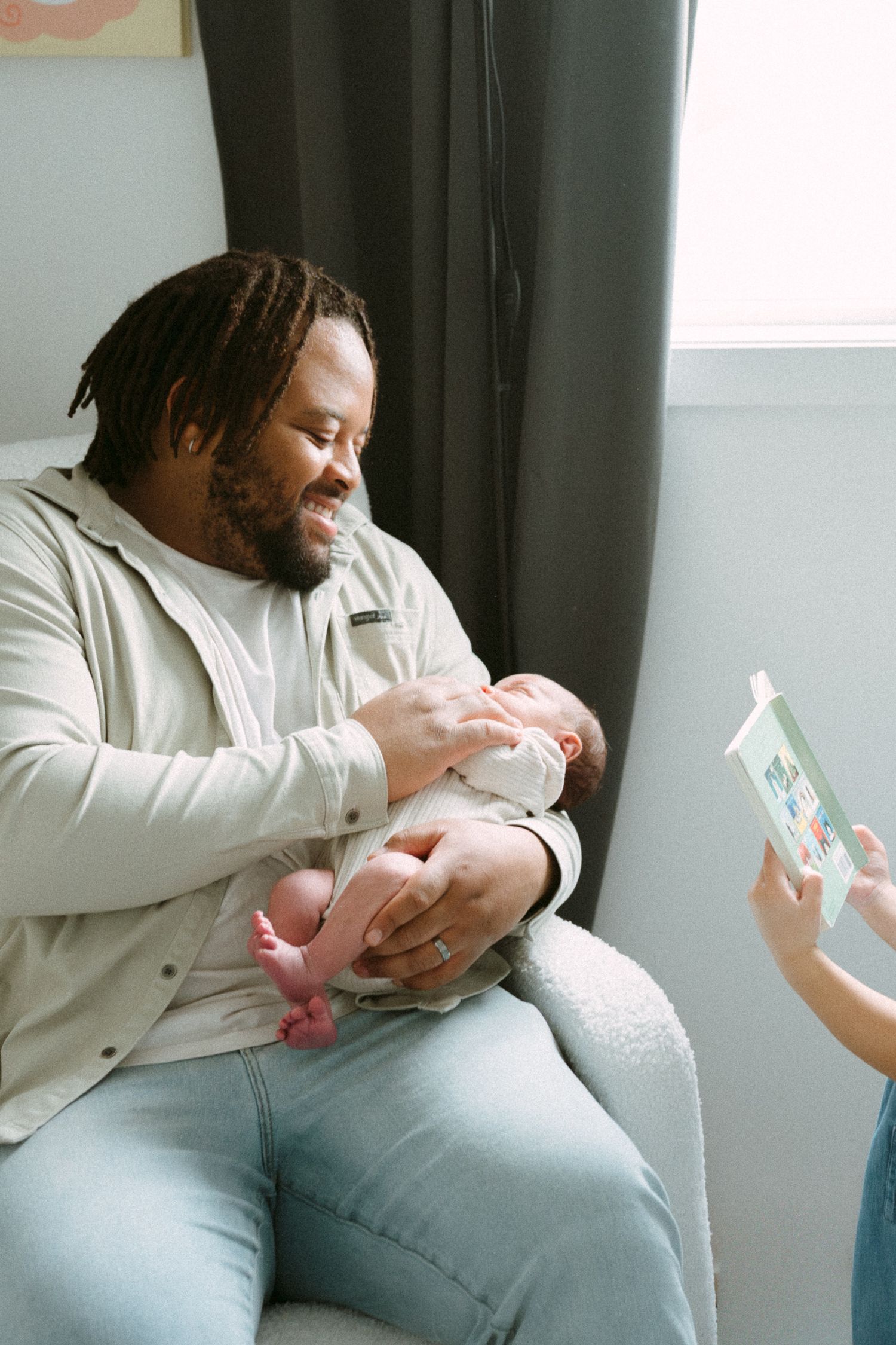 Happy Black father with dreadlocks smiles while cradling newborn baby in a cozy nursery chair.