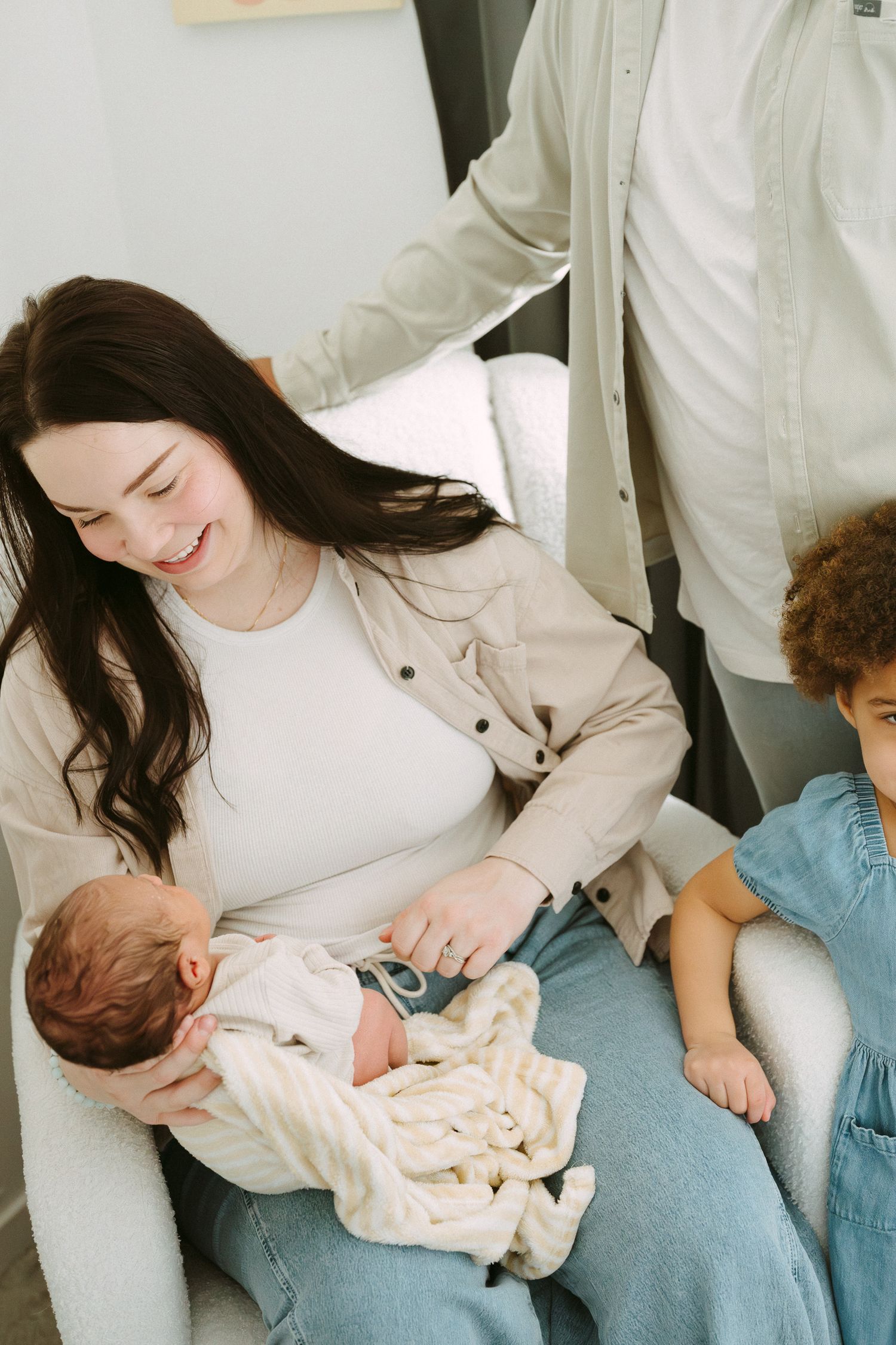 Smiling mother holds newborn baby in white blanket while sitting in chair with toddler beside her.