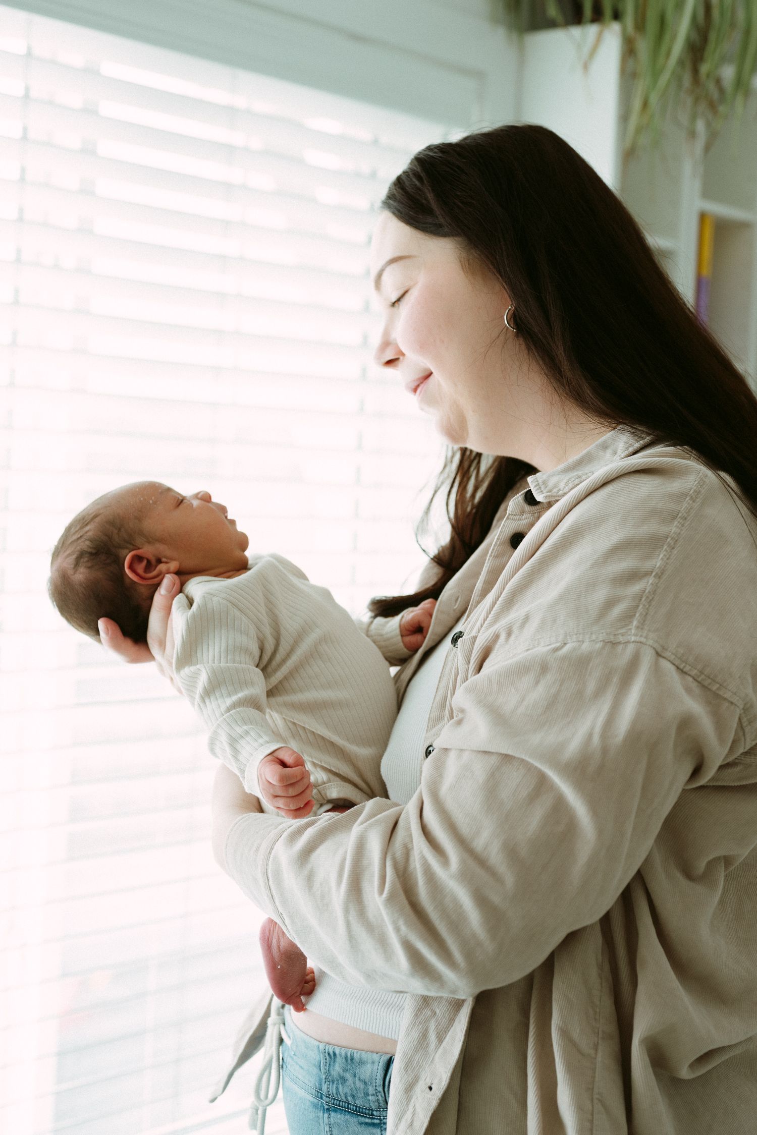 A young mother smiles tenderly while cradling her newborn baby near a bright window with white blinds.
