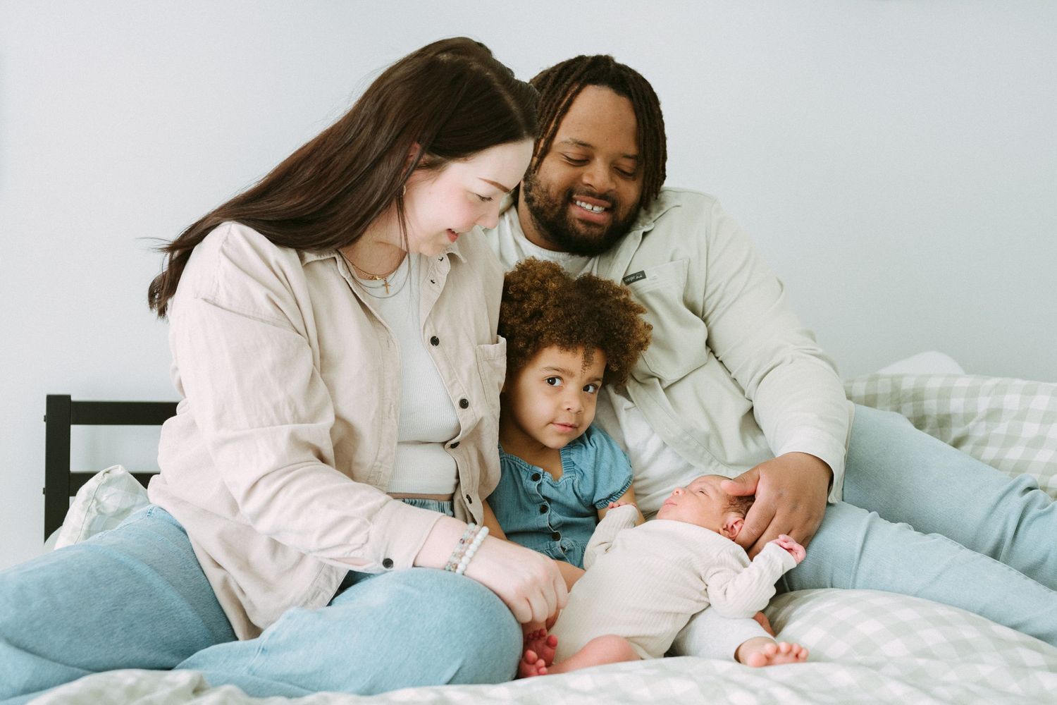 Multiracial family with toddler and newborn sitting together on bed.