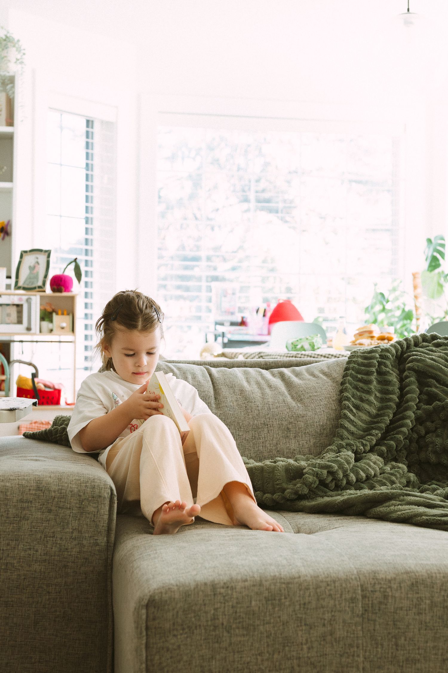 A child lies comfortably reading on a gray couch with a knitted blanket.