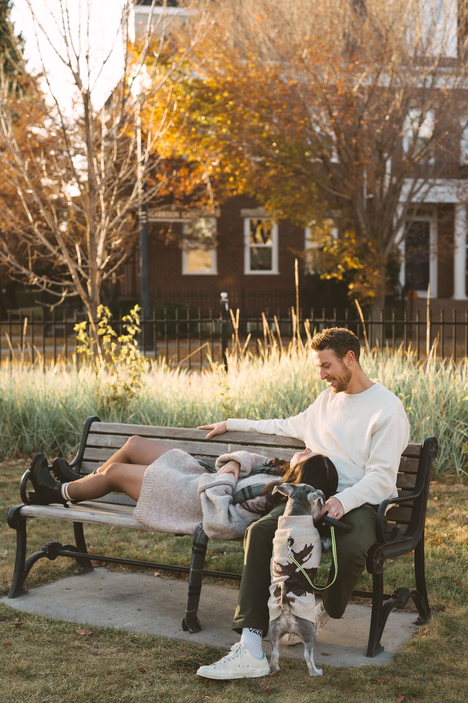 A couple relaxes on a park bench with their white dog on an autumn afternoon.