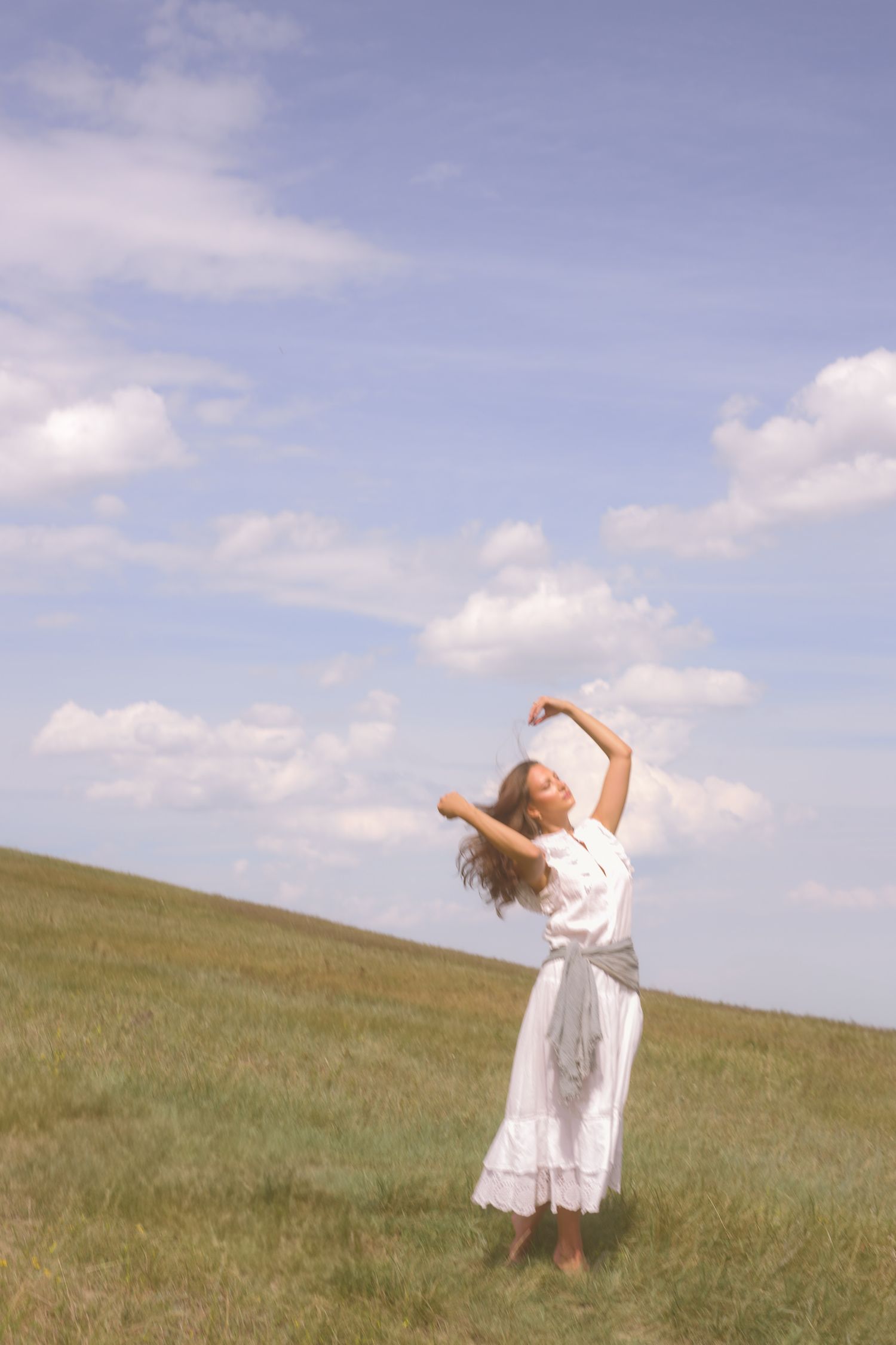 A graceful figure in white dances freely on a grassy hillside under blue skies.