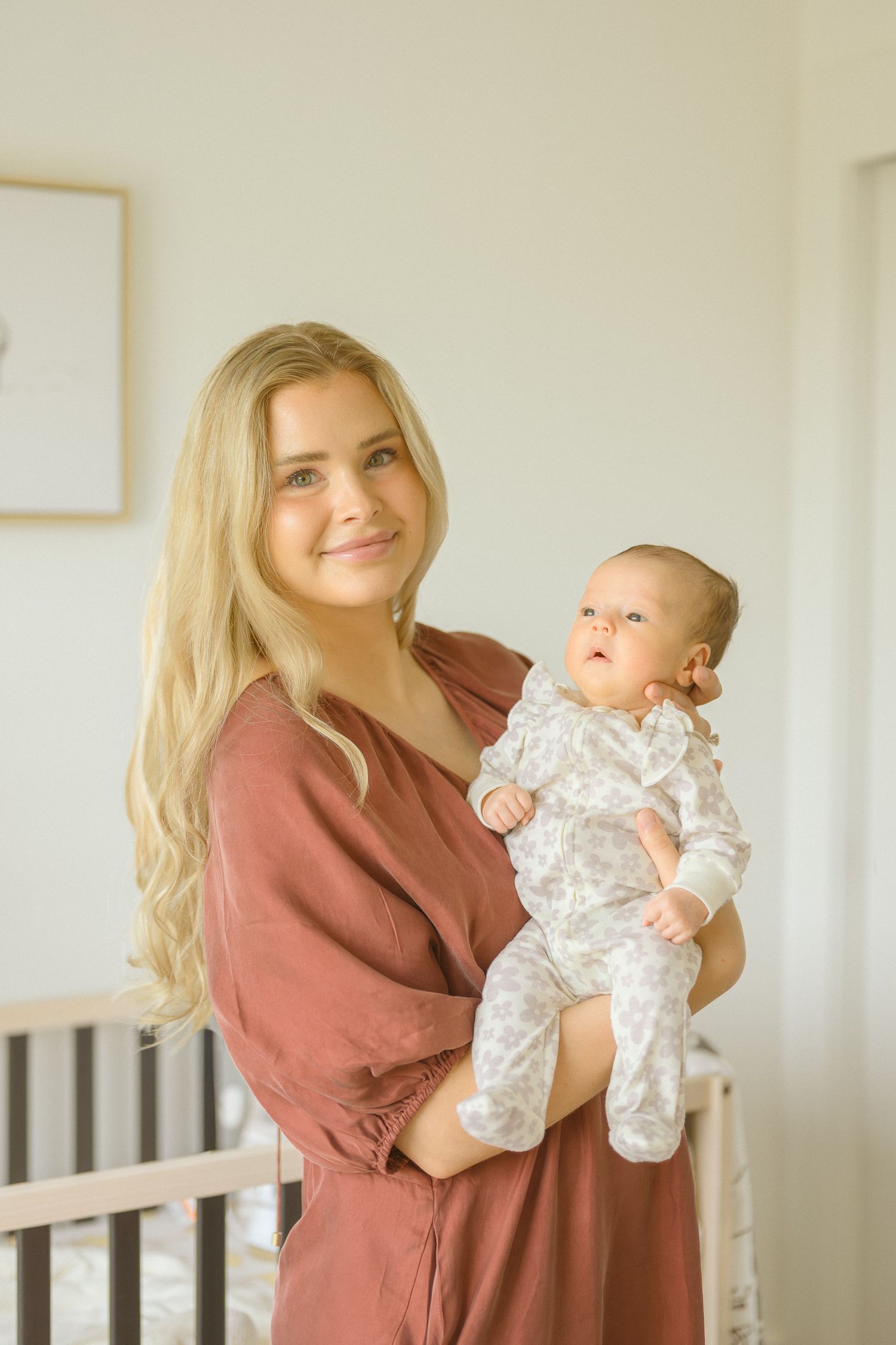 A woman in a mauve dress holds a baby wearing white floral pajamas in a minimalist nursery setting.