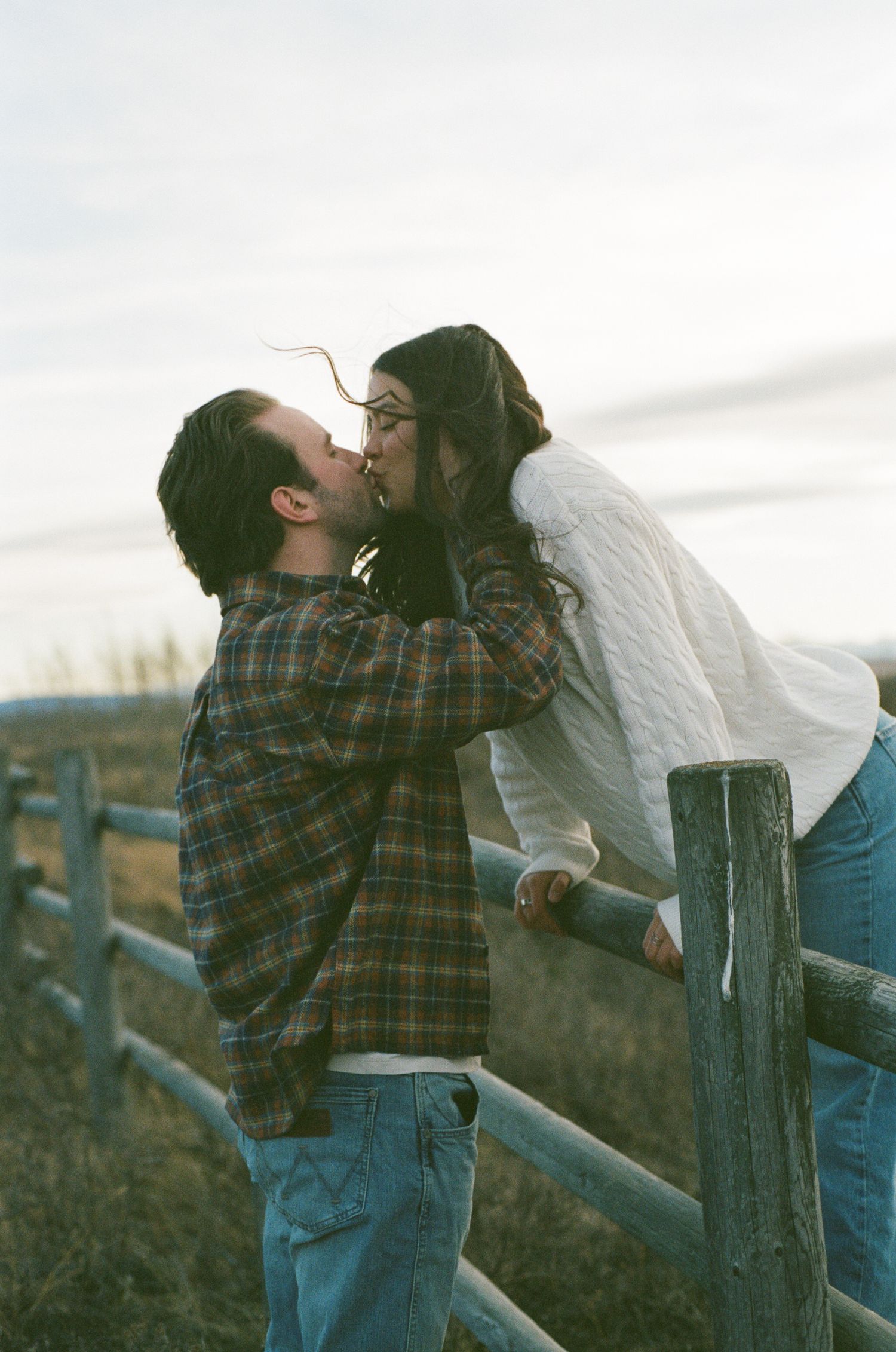 A couple shares an intimate moment while leaning against a wooden fence in the countryside.