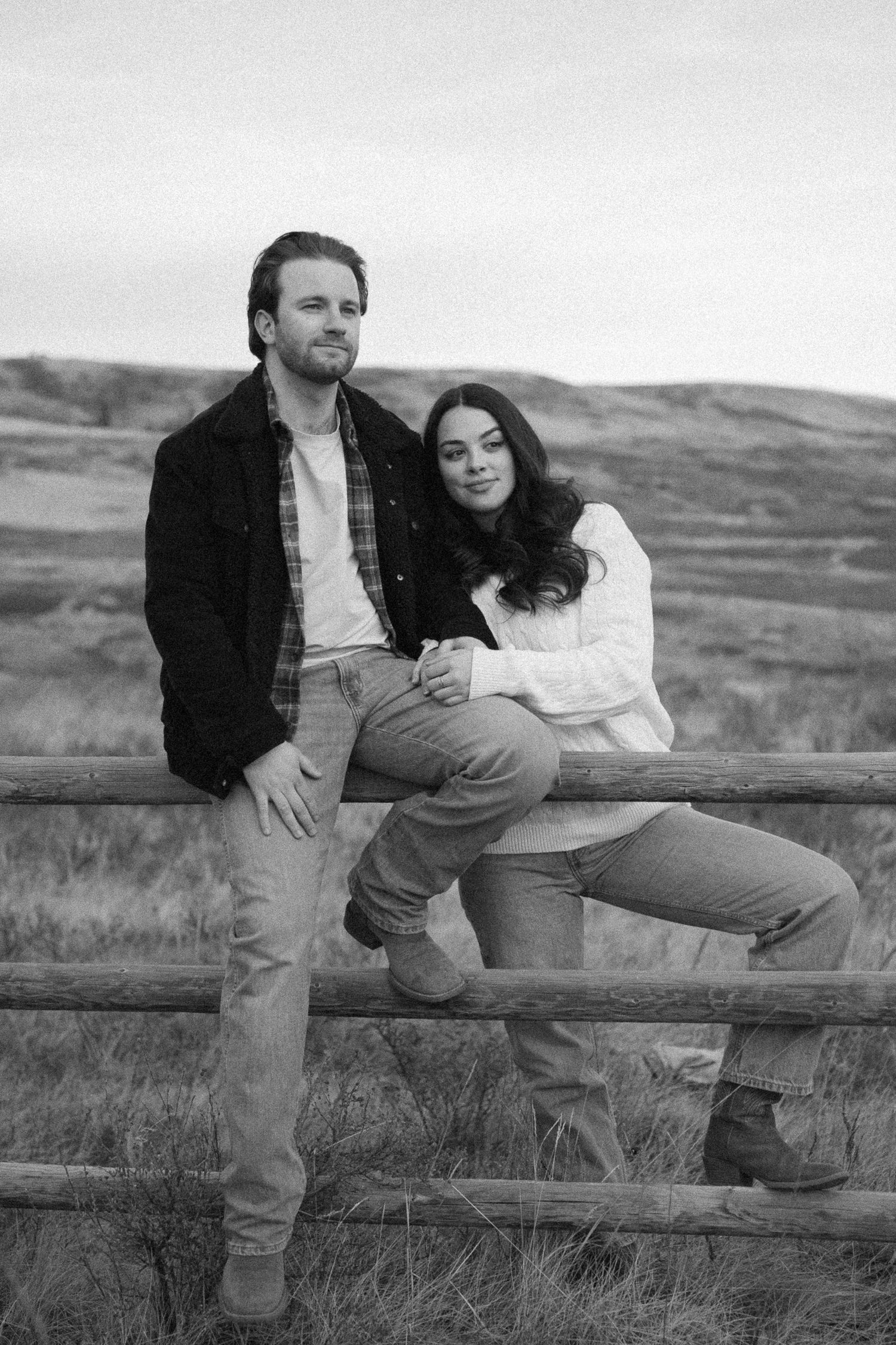 Black and white photo of a couple sitting together on a rustic wooden fence in a rural field.