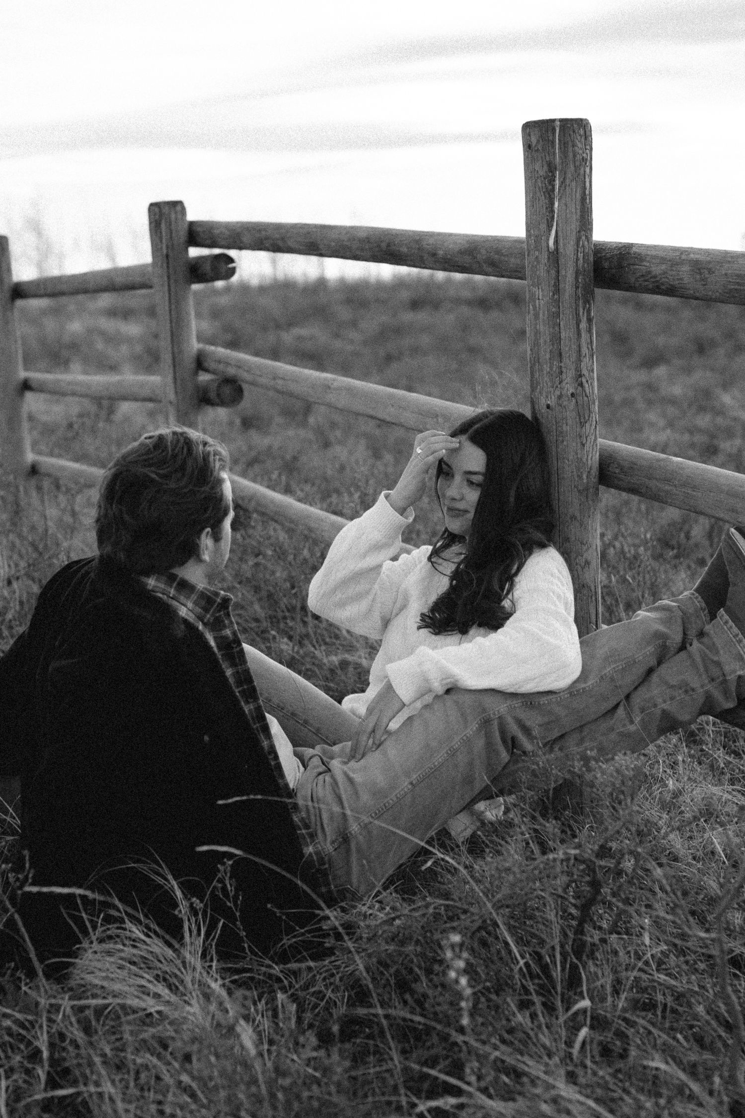 Two people sit together in tall grass near a wooden fence in a black and white vintage photograph.