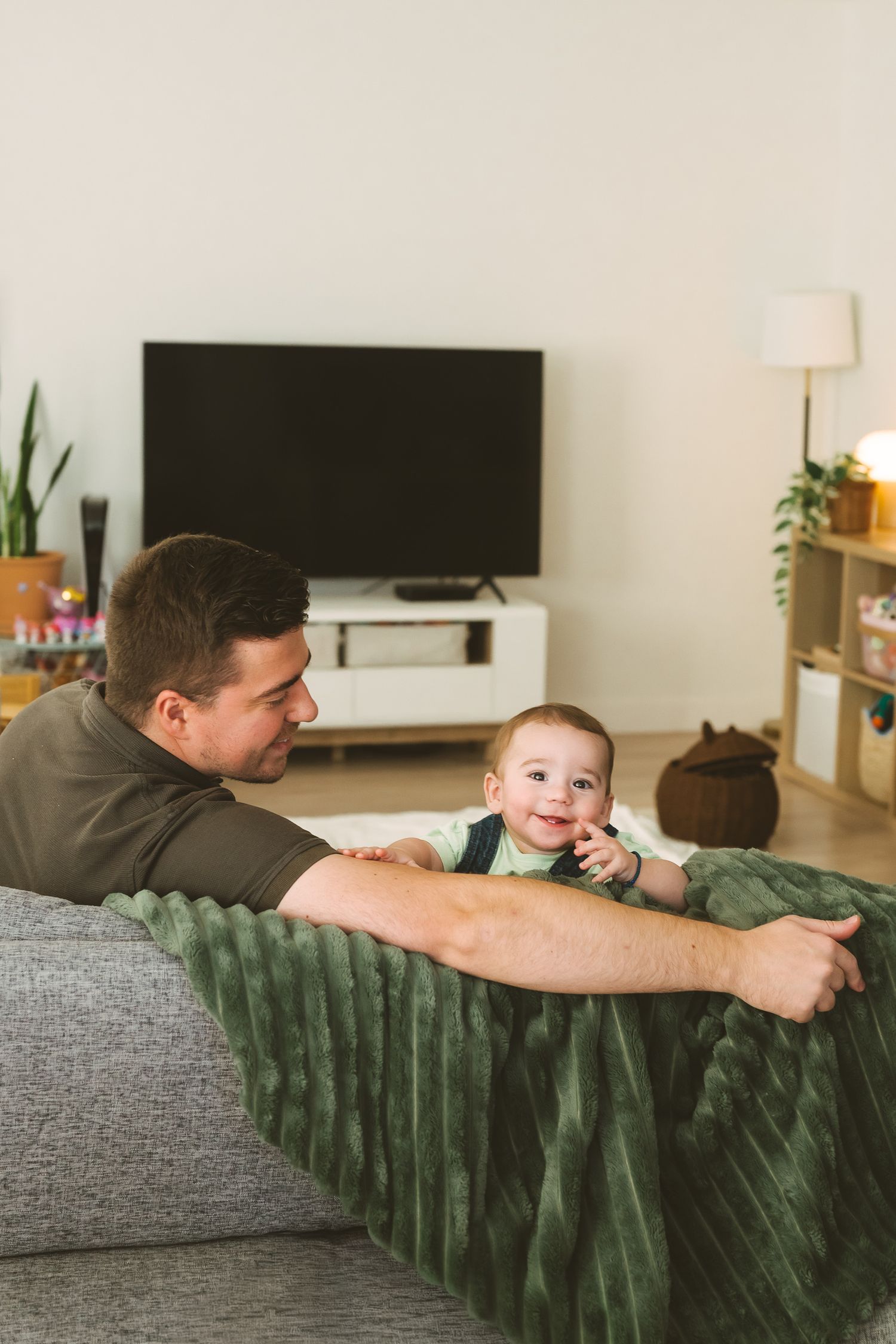 Relaxed family moment on grey couch with TV and houseplants in background.