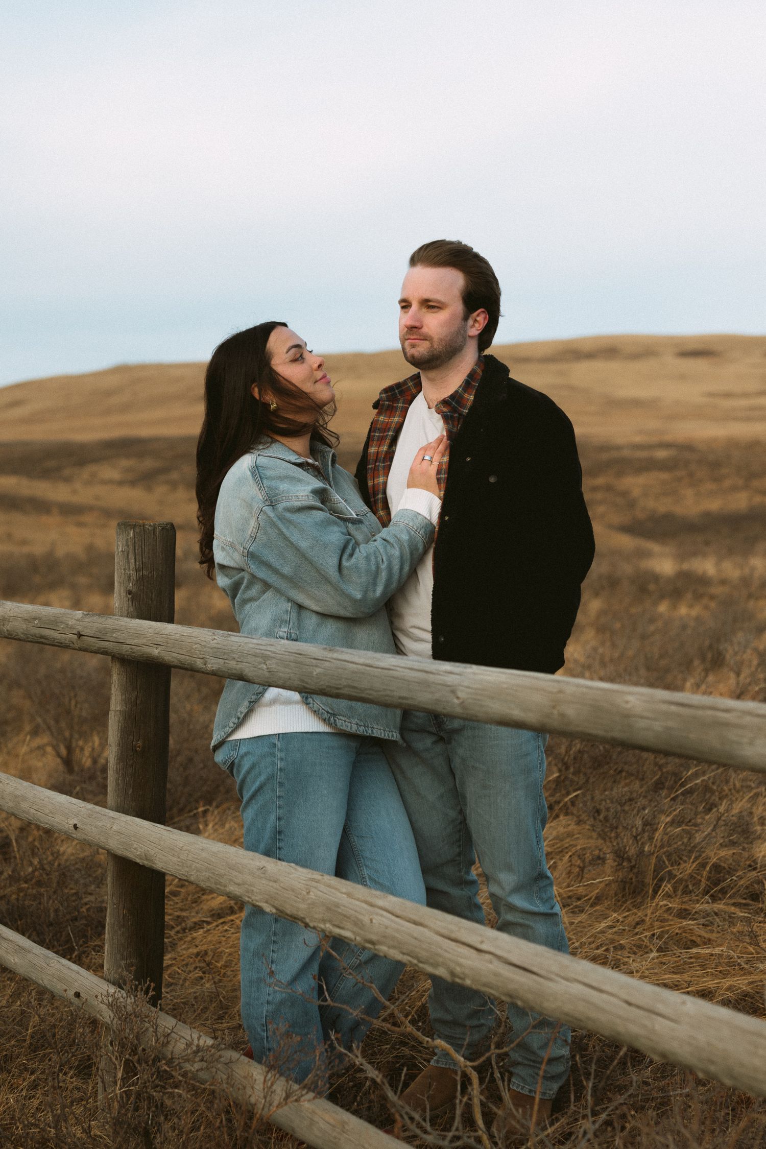 A romantic couple shares an intimate moment while leaning against a wooden fence in a rural setting.