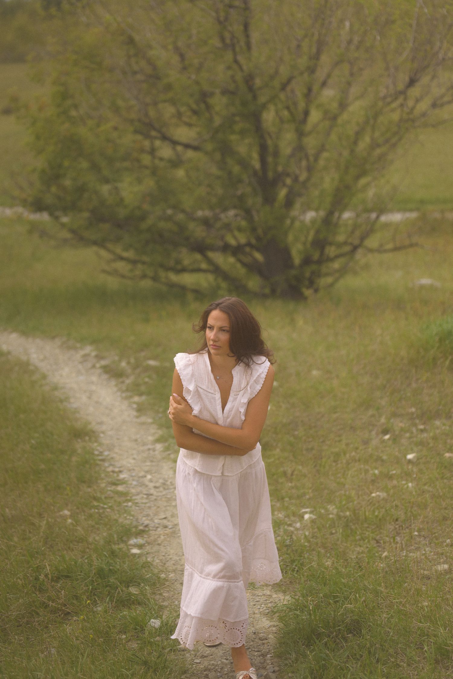 Someone in a white dress walks along a winding dirt path through spring meadow grass.