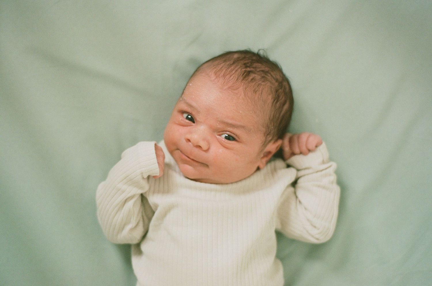 A newborn baby in a cream onesie lies on a mint green blanket with eyes open.