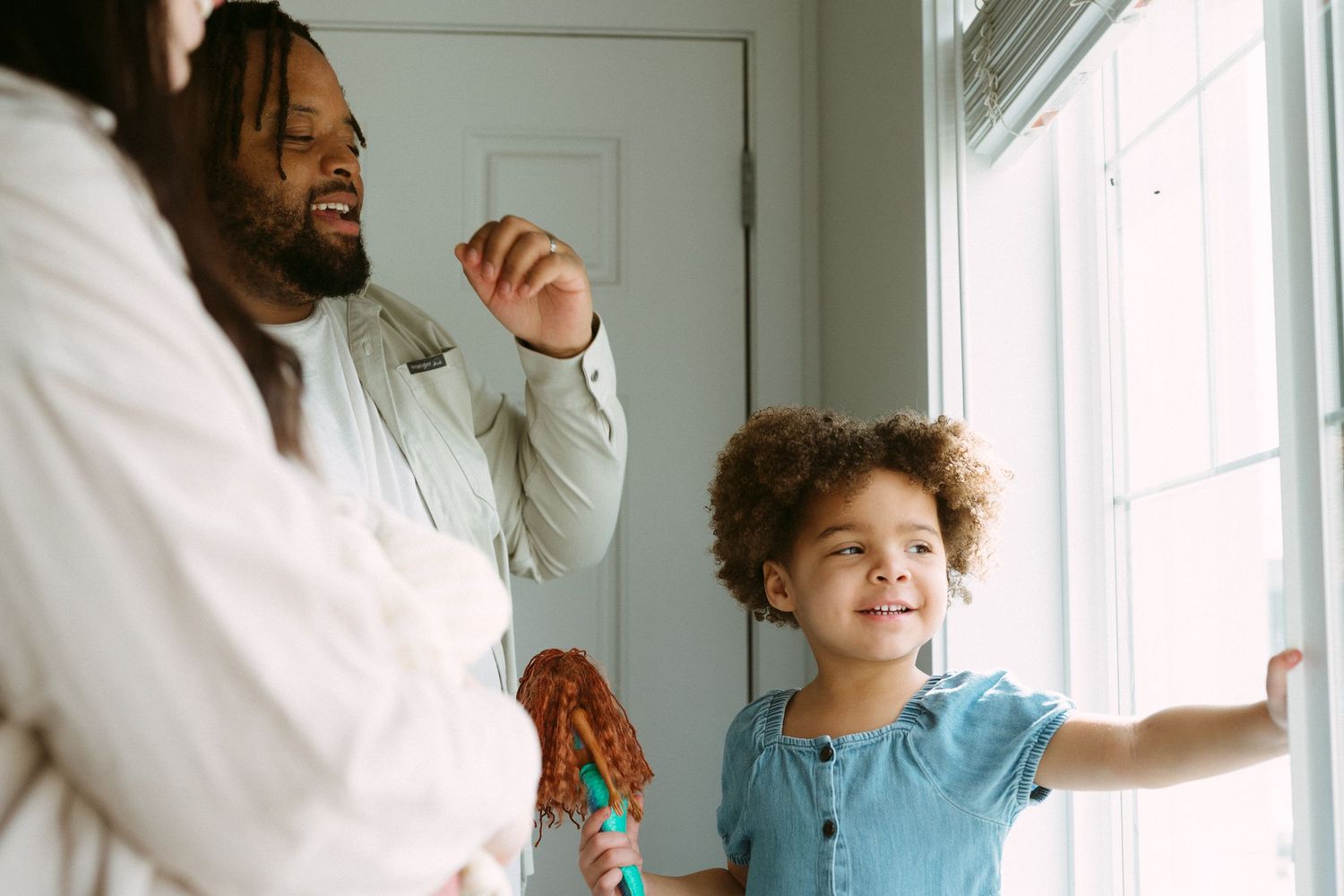 A father and young girl with curly hair smile and play near a bright window in a cozy home setting.