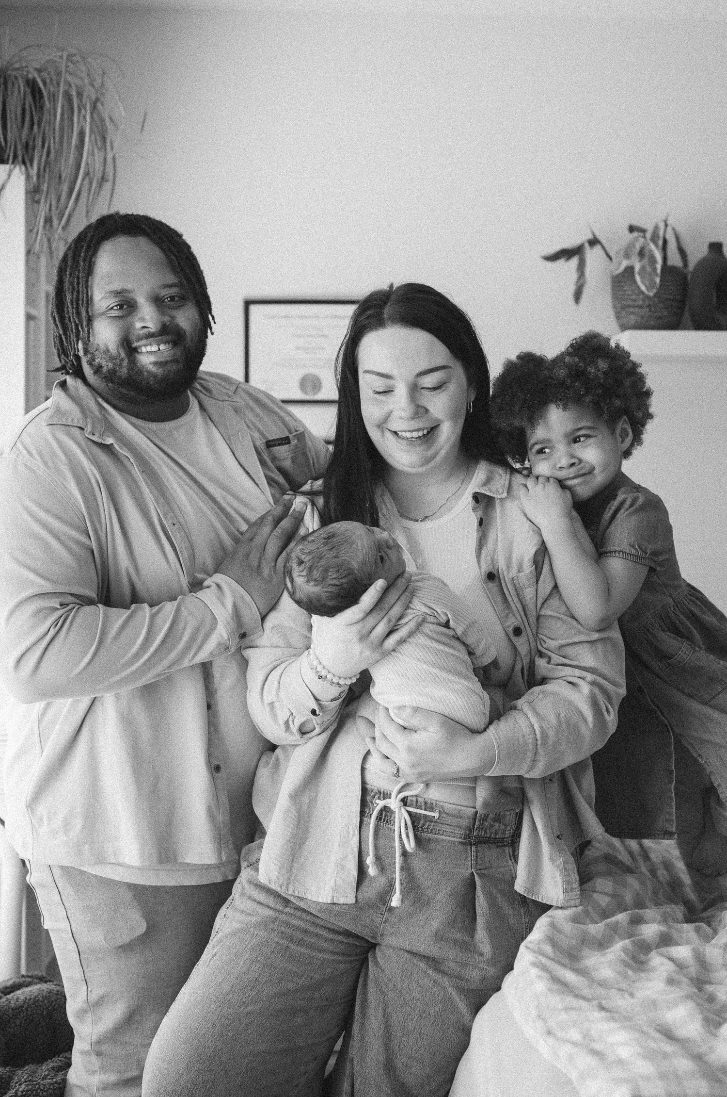 Black and white photo of a smiling family with a newborn baby and young child posing together indoors.