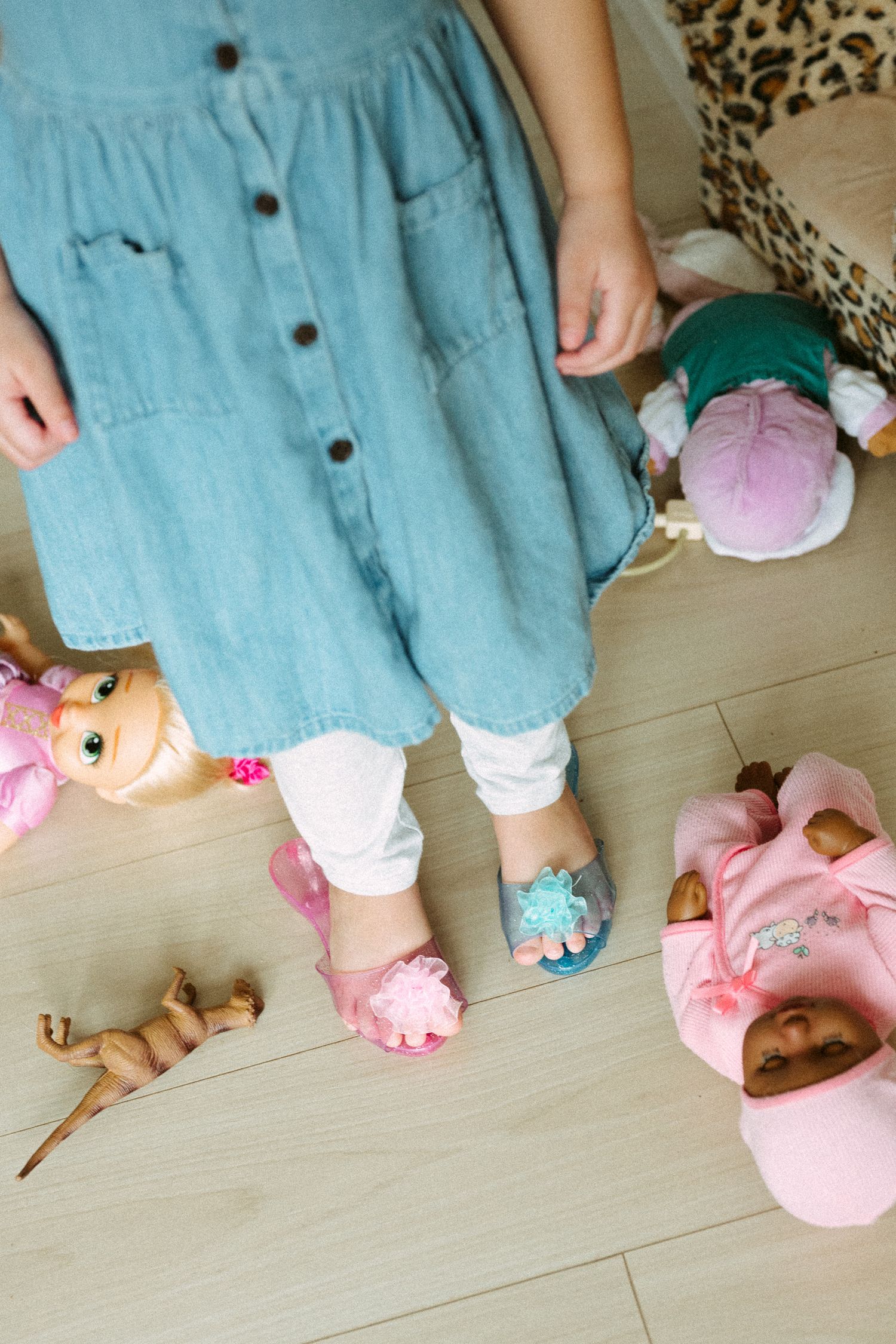 Young girl in denim dress and sandals standing on wood floor surrounded by baby dolls and toys.