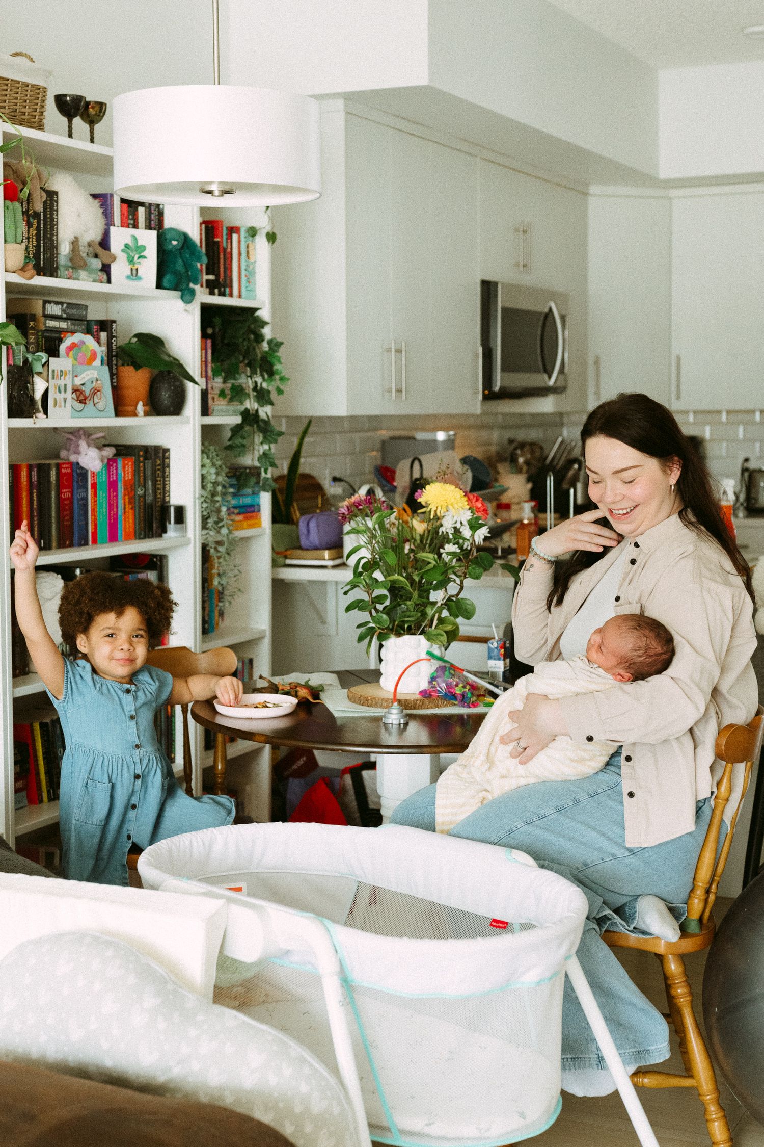 A smiling mother holds her newborn baby while sitting in a rocking chair as her young toddler plays nearby in a cozy home.