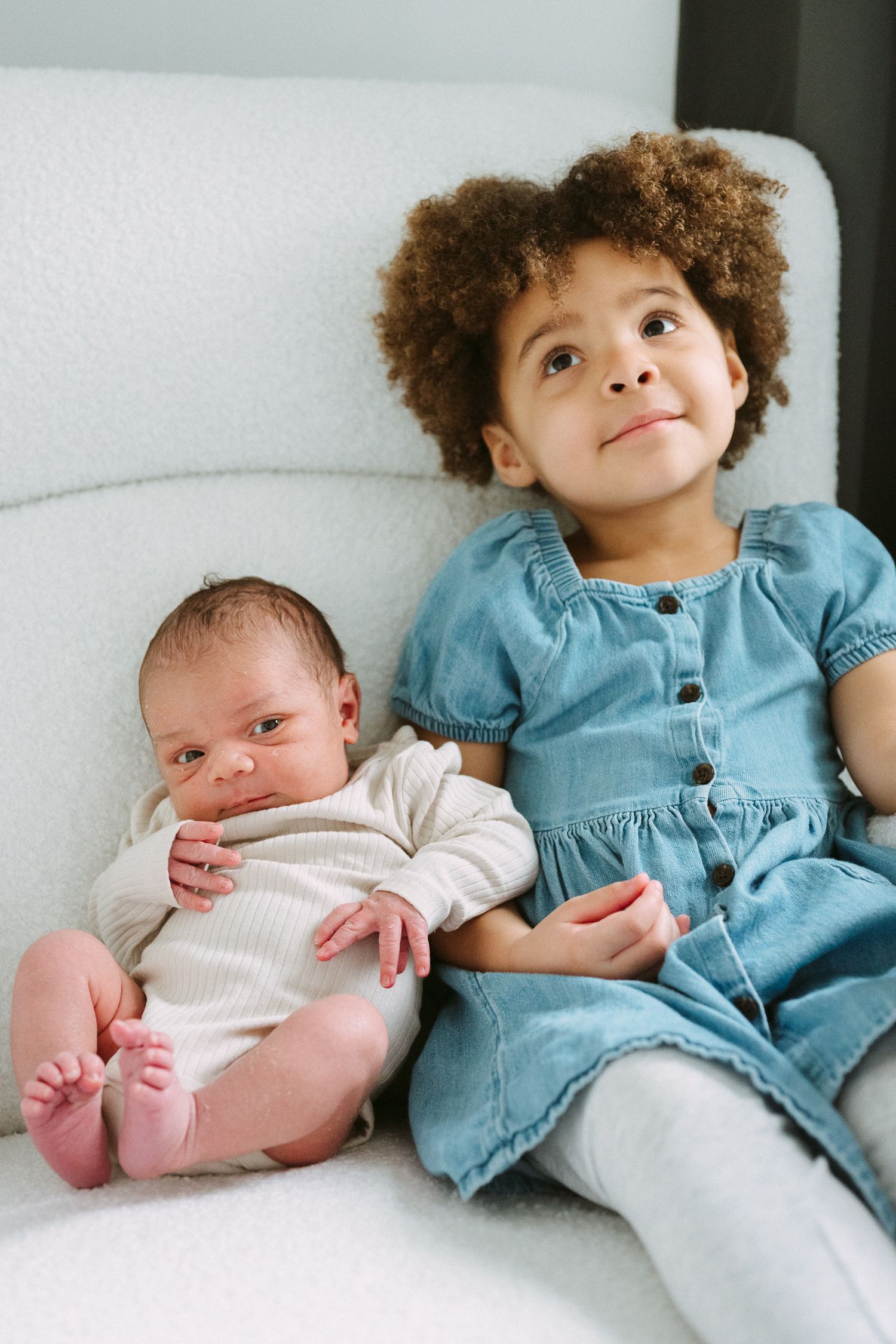 Curly-haired toddler girl in denim dress sits in white chair holding newborn baby sibling while looking up.