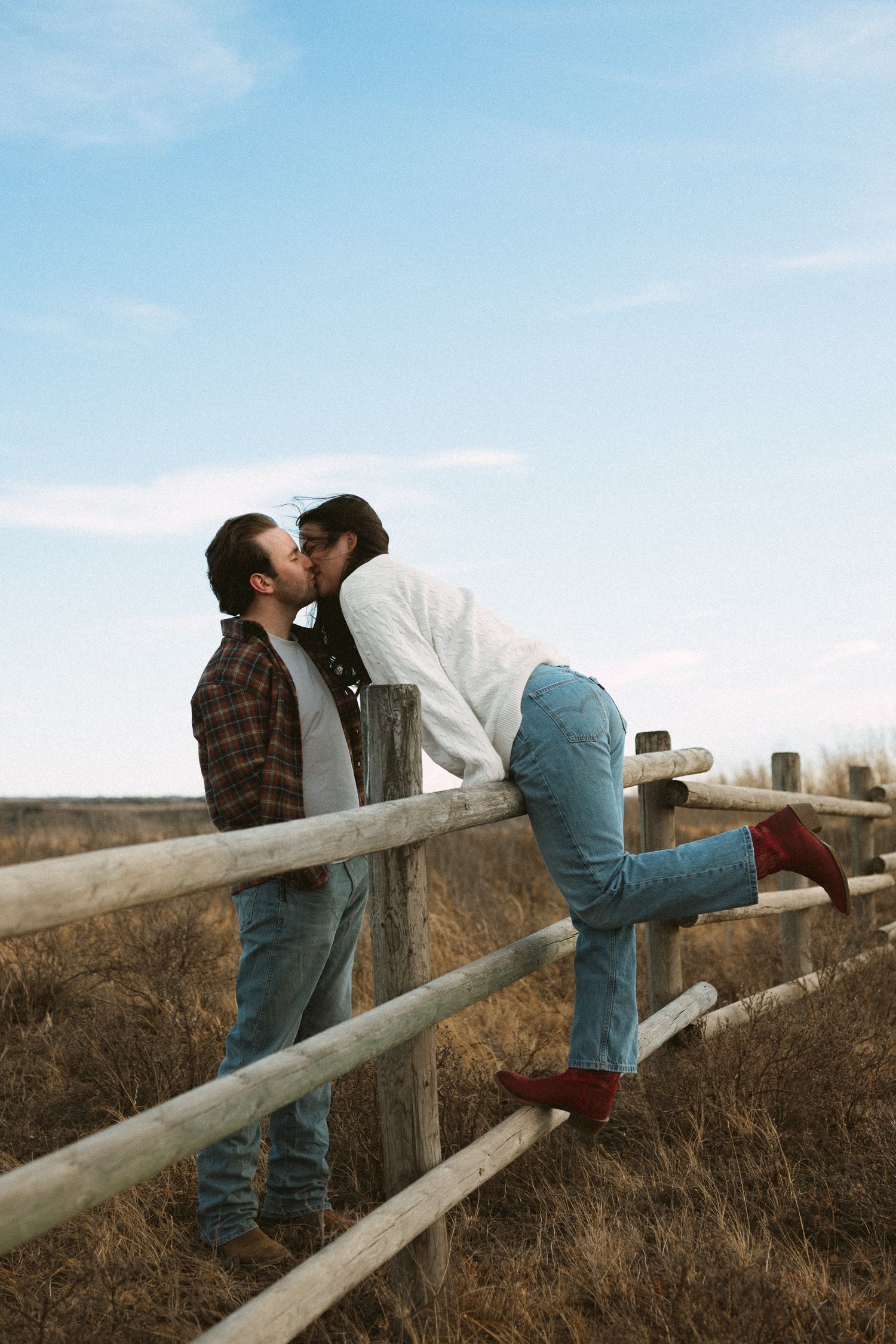 A couple shares a romantic moment leaning against a wooden fence rail in a rustic outdoor setting.