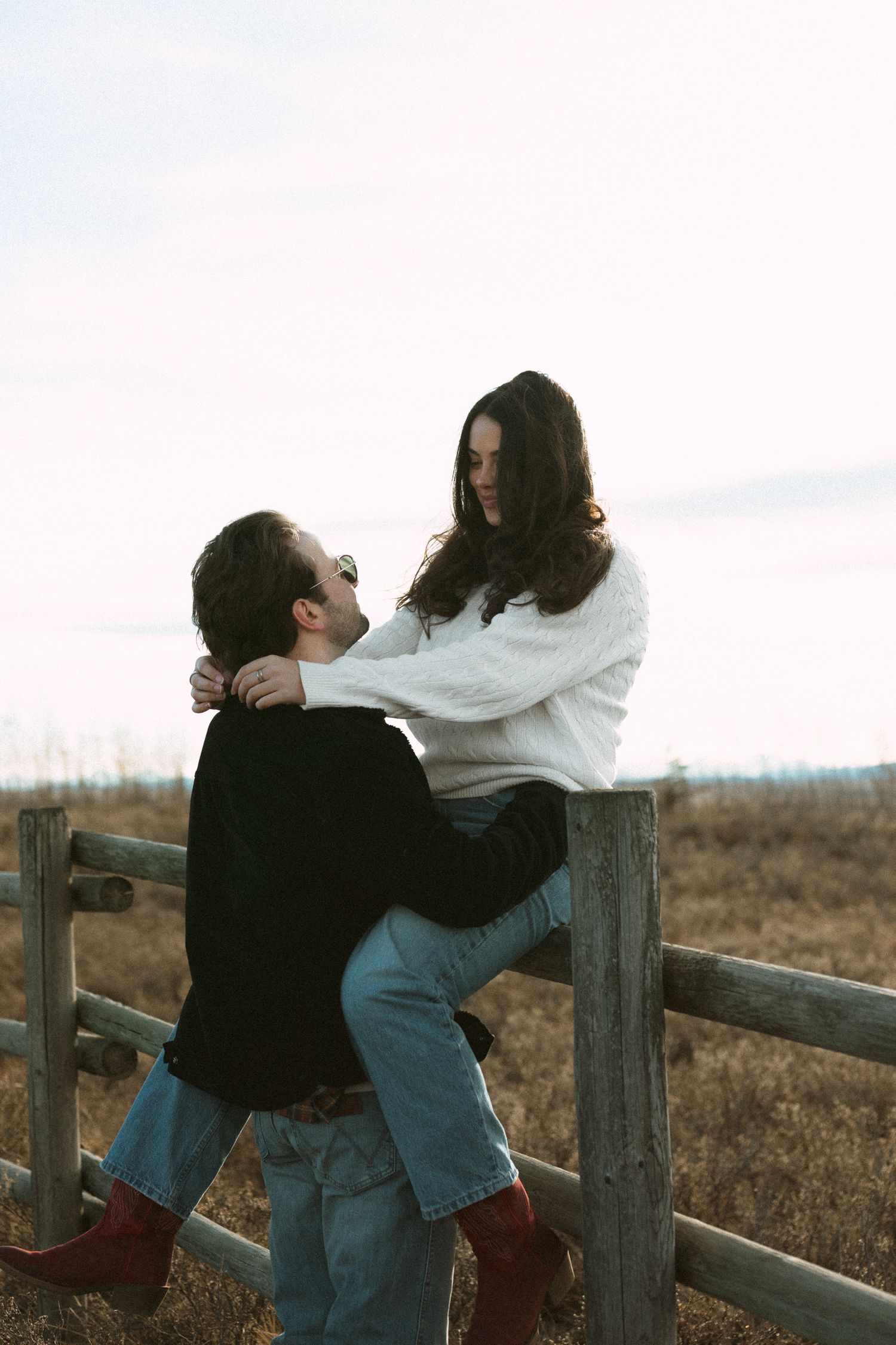 Couple embracing against rustic wooden fence at sunset in countryside setting.