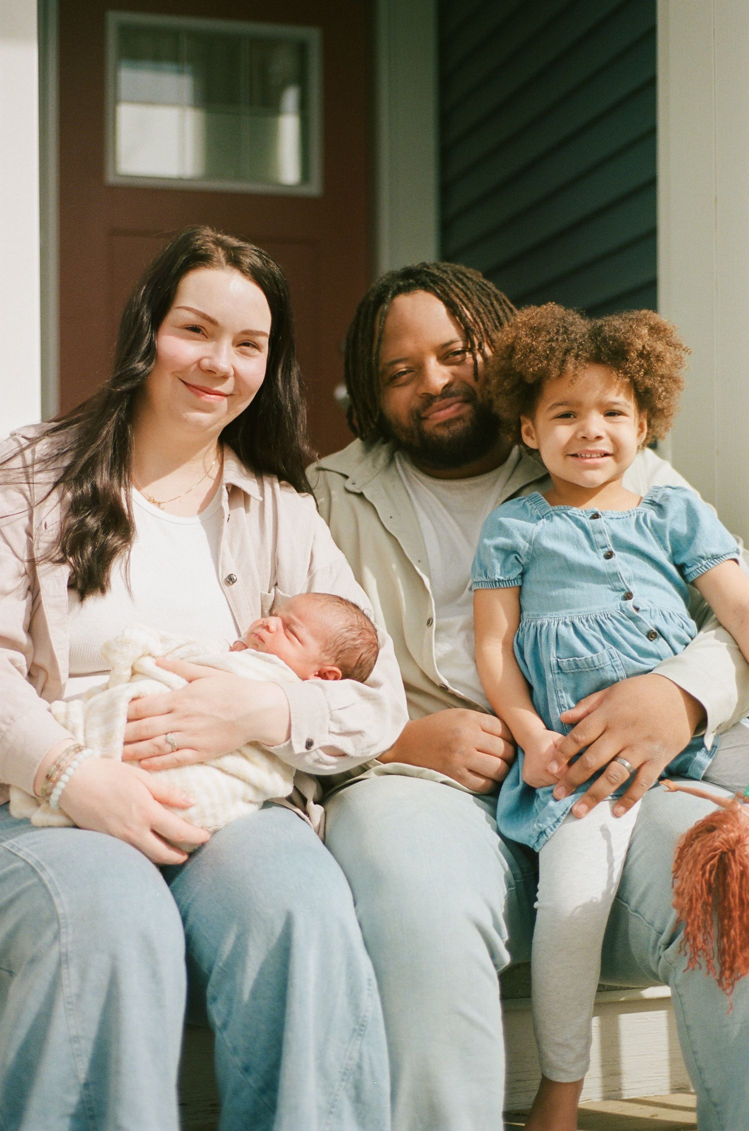 Happy family of four sitting on porch steps with newborn baby and toddler girl.