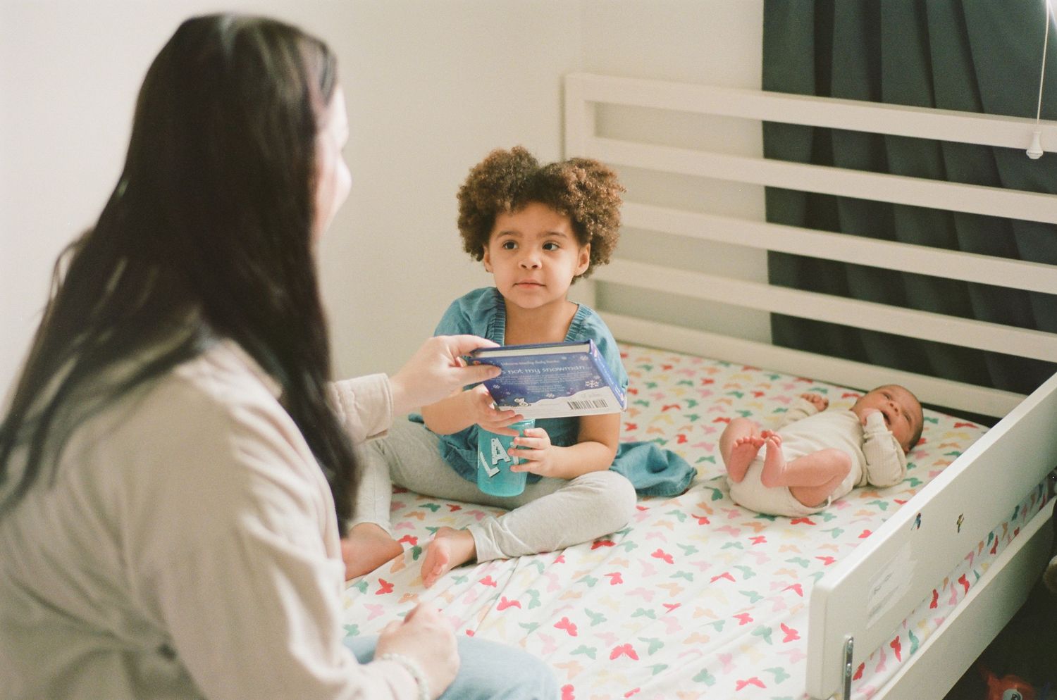 A woman sits on a bed talking to a young curly-haired child holding a tablet in a bright bedroom.