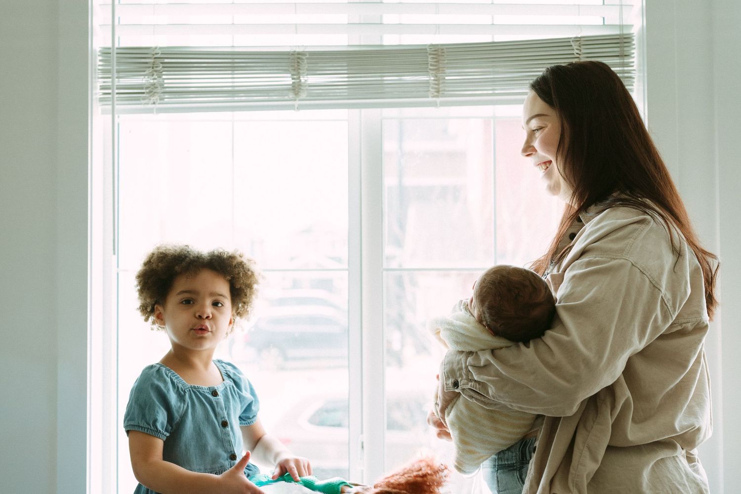 A mother holds her newborn baby while smiling at her young curly-haired child standing by a bright window.