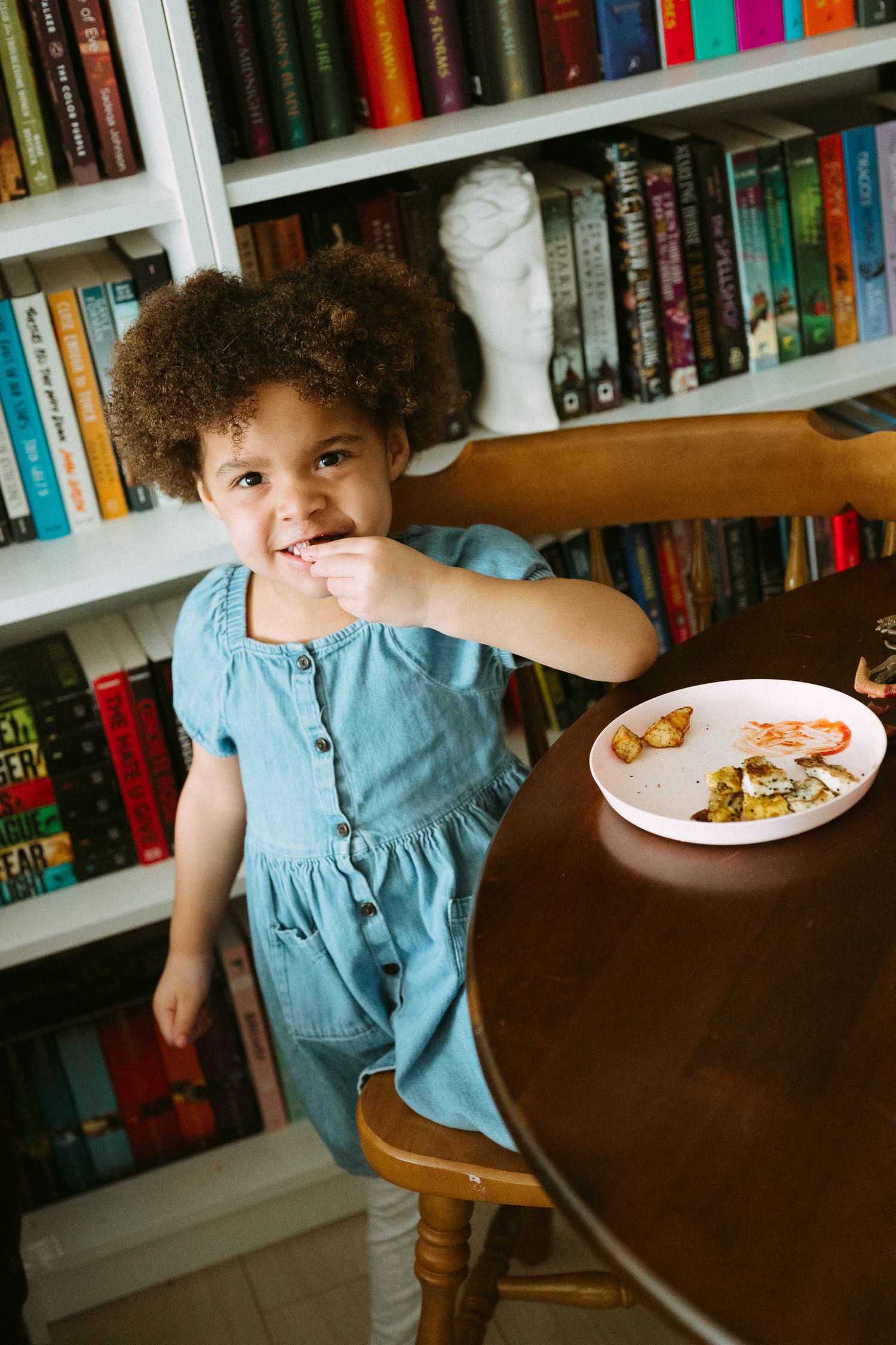 A young girl with curly hair in a blue denim dress eating at a wooden table near colorful bookshelves.