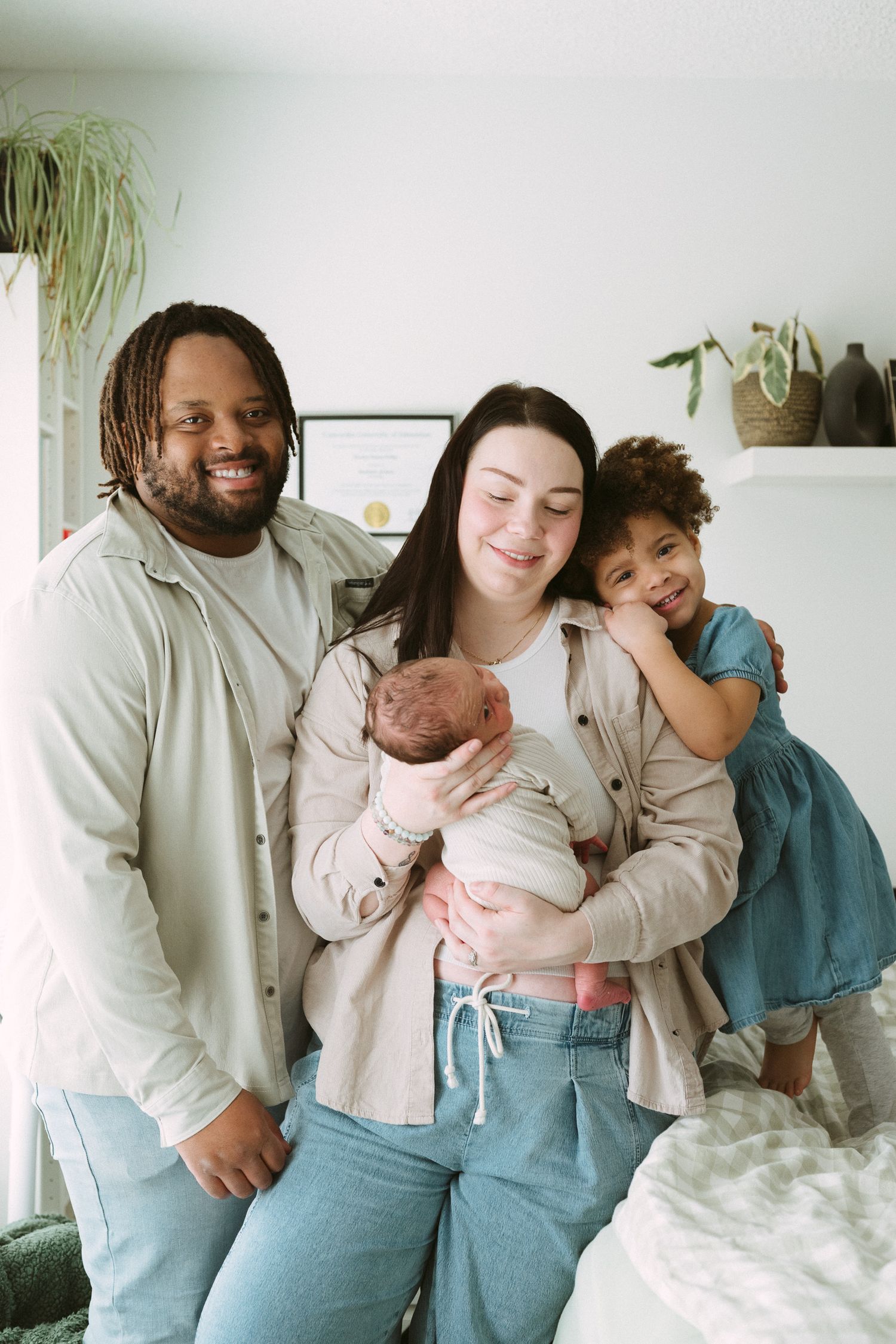 Happy interracial family of four posing indoors, parents holding newborn baby with young daughter smiling beside them.