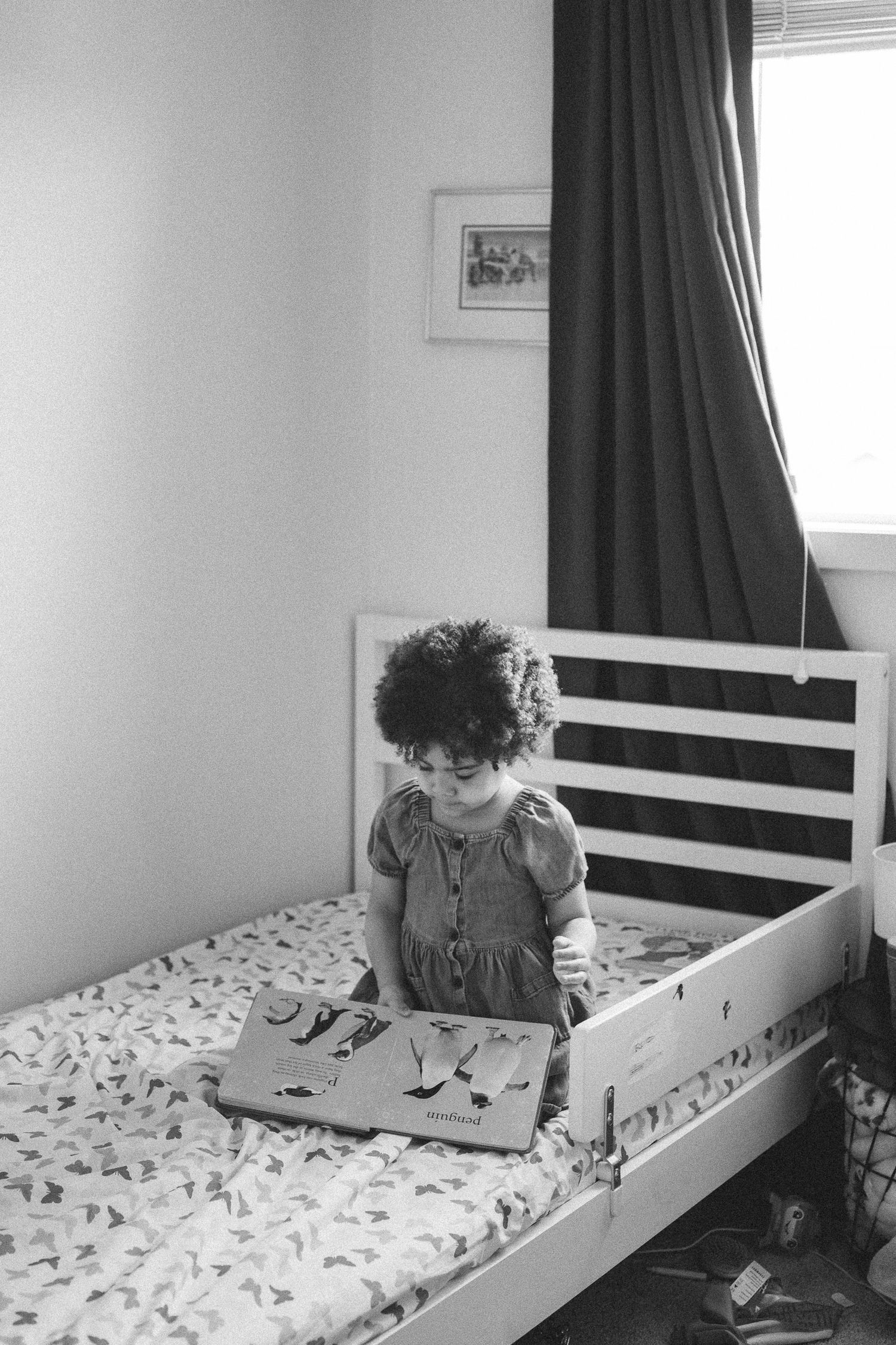 Black and white photo of a young girl with curly hair sitting on a bed, looking at an open book in a cozy bedroom.